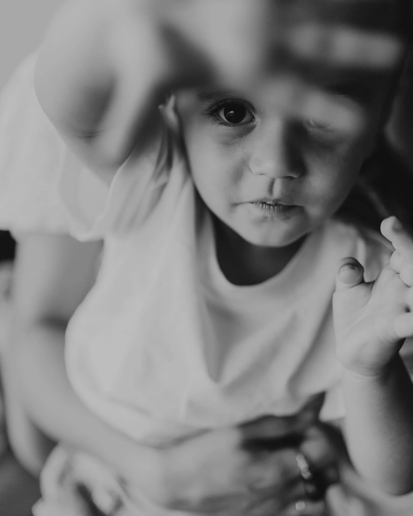 Close-up black-and-white photo of a young child with wide eyes looking into the camera, holding her hand near her face.