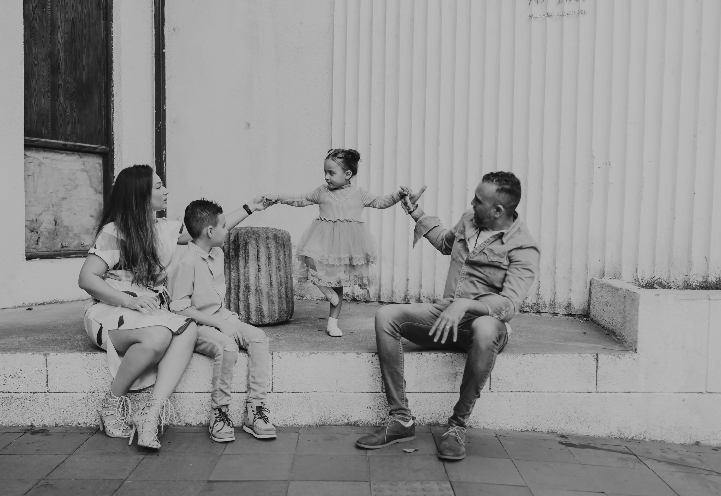 A black and white image of a family sitting on a sidewalk outside, with two children and two adults. The young girl is standing and dancing with arms extended, holding the hands of a woman on her left and a man on her right. The woman is sitting and 