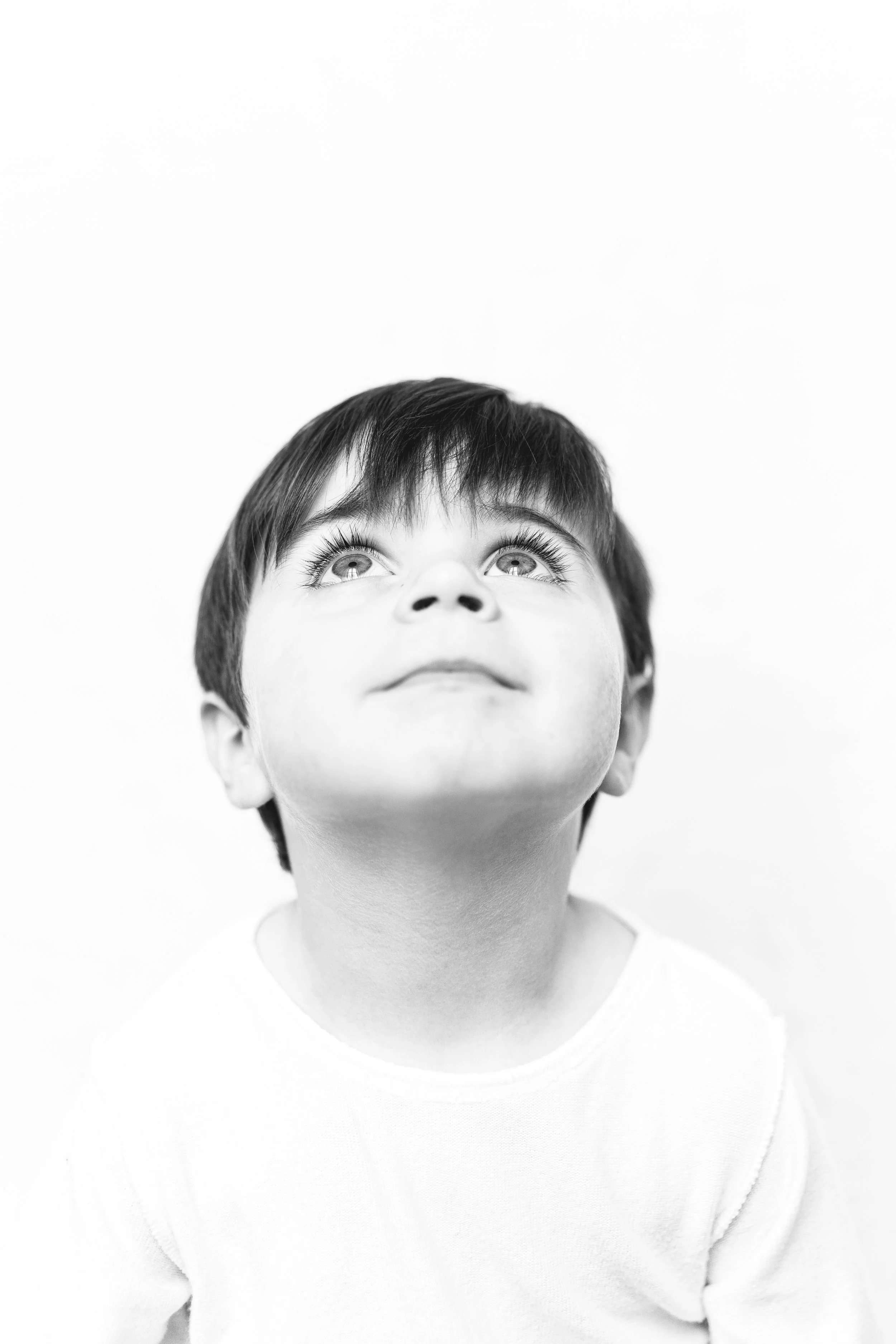 A young boy with dark hair looking upward with a curious and thoughtful expression, photographed in black and white against a plain background.
