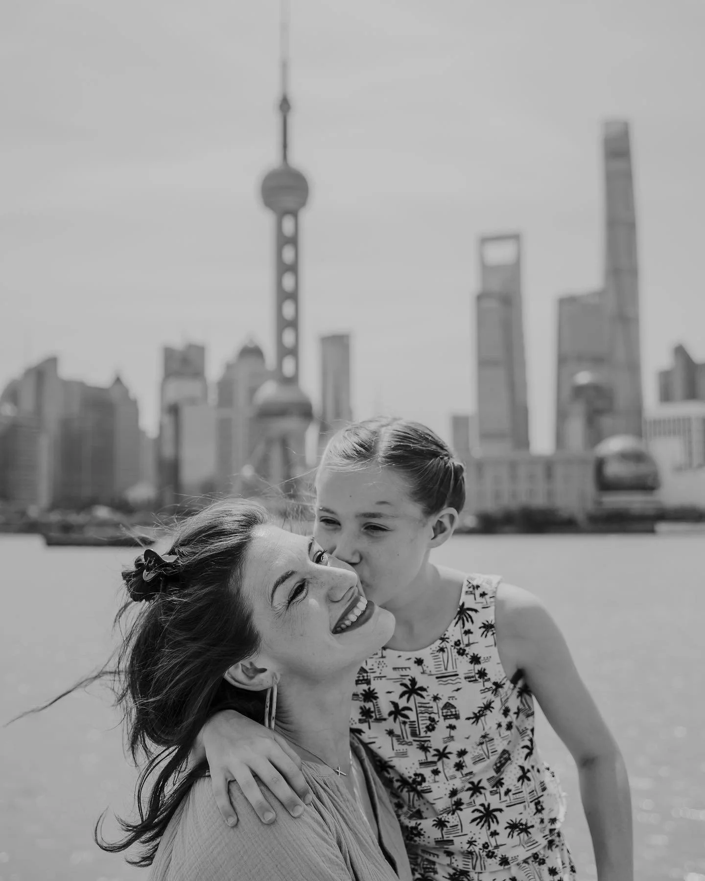 A woman and a girl, likely mother and daughter, sharing a kiss near a body of water with a city skyline and tall buildings in the background.
