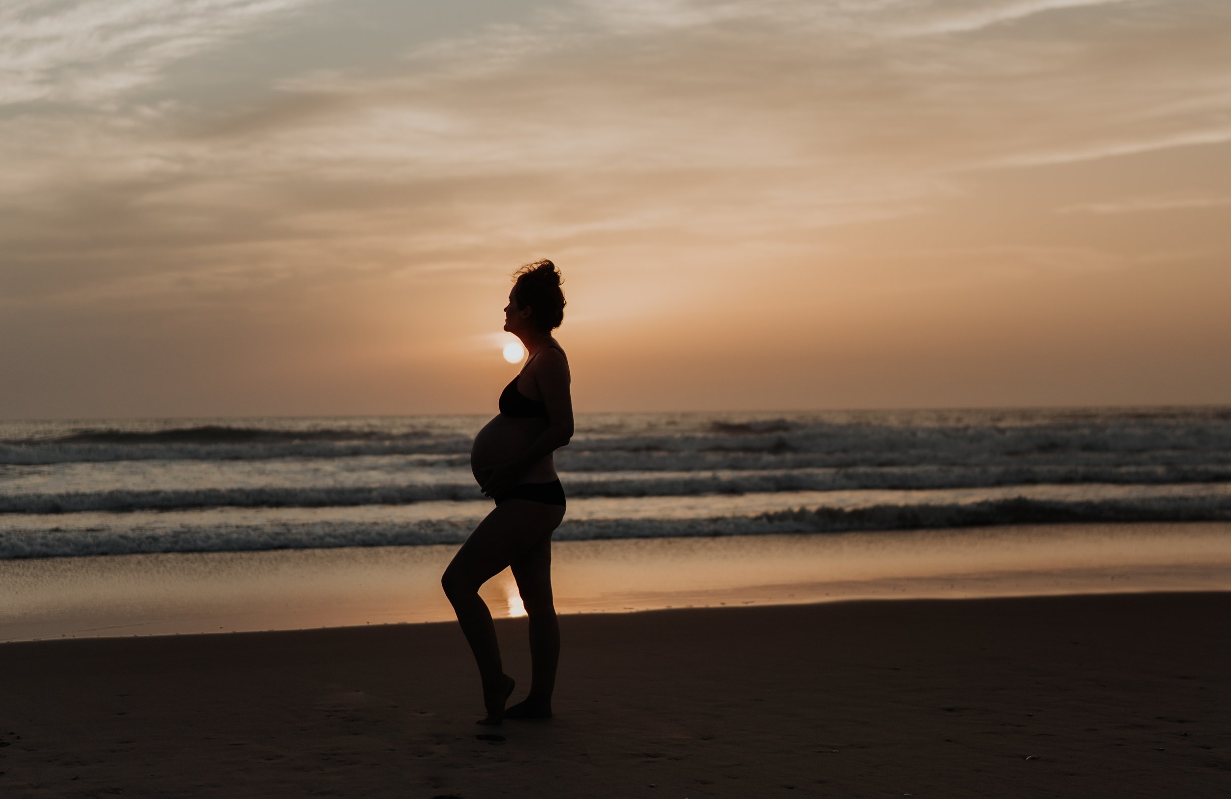 Silhouette of a pregnant woman standing on the beach at sunset.