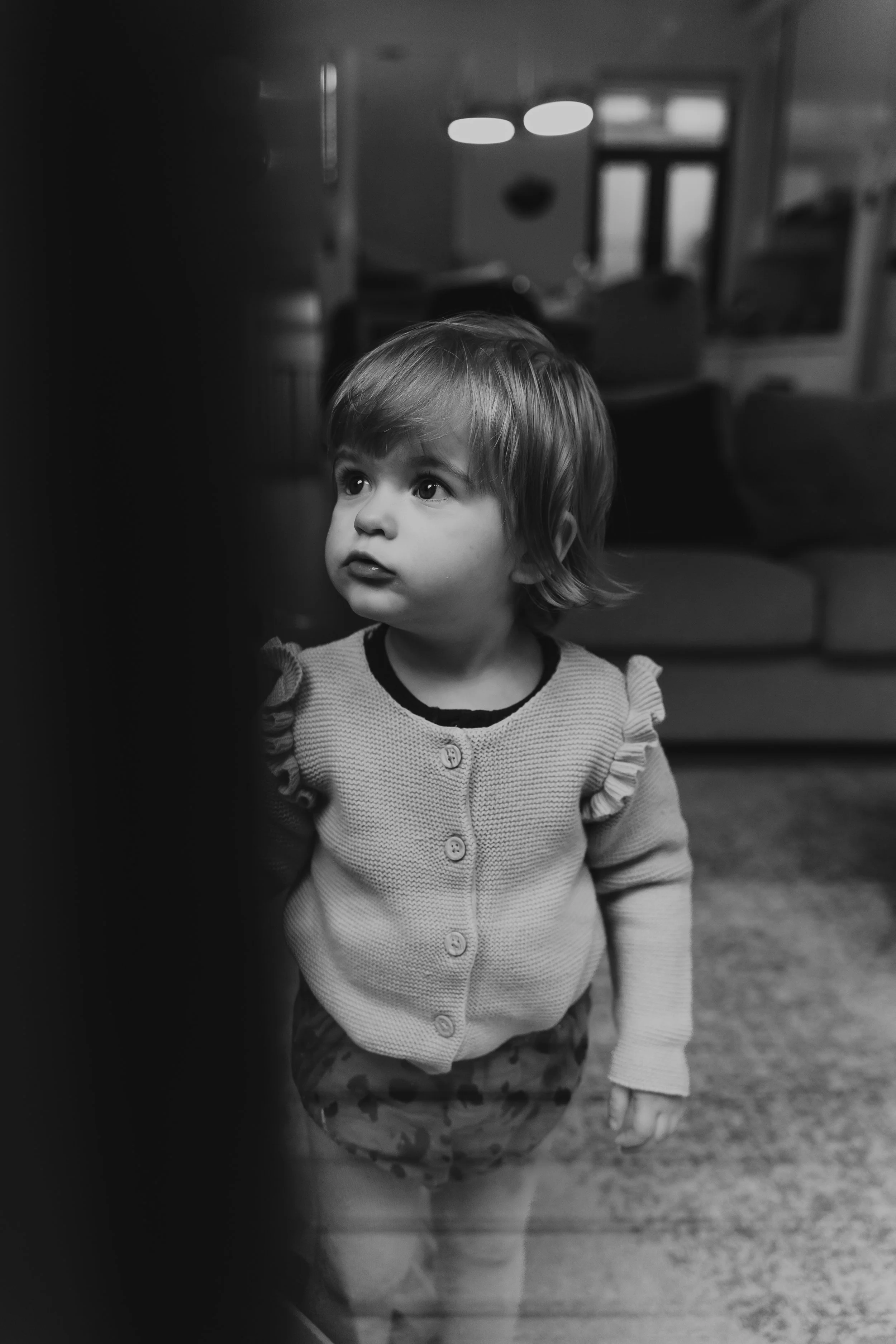 A young girl with short hair wearing a sweater and patterned shorts, standing indoors, looking intently to the side.