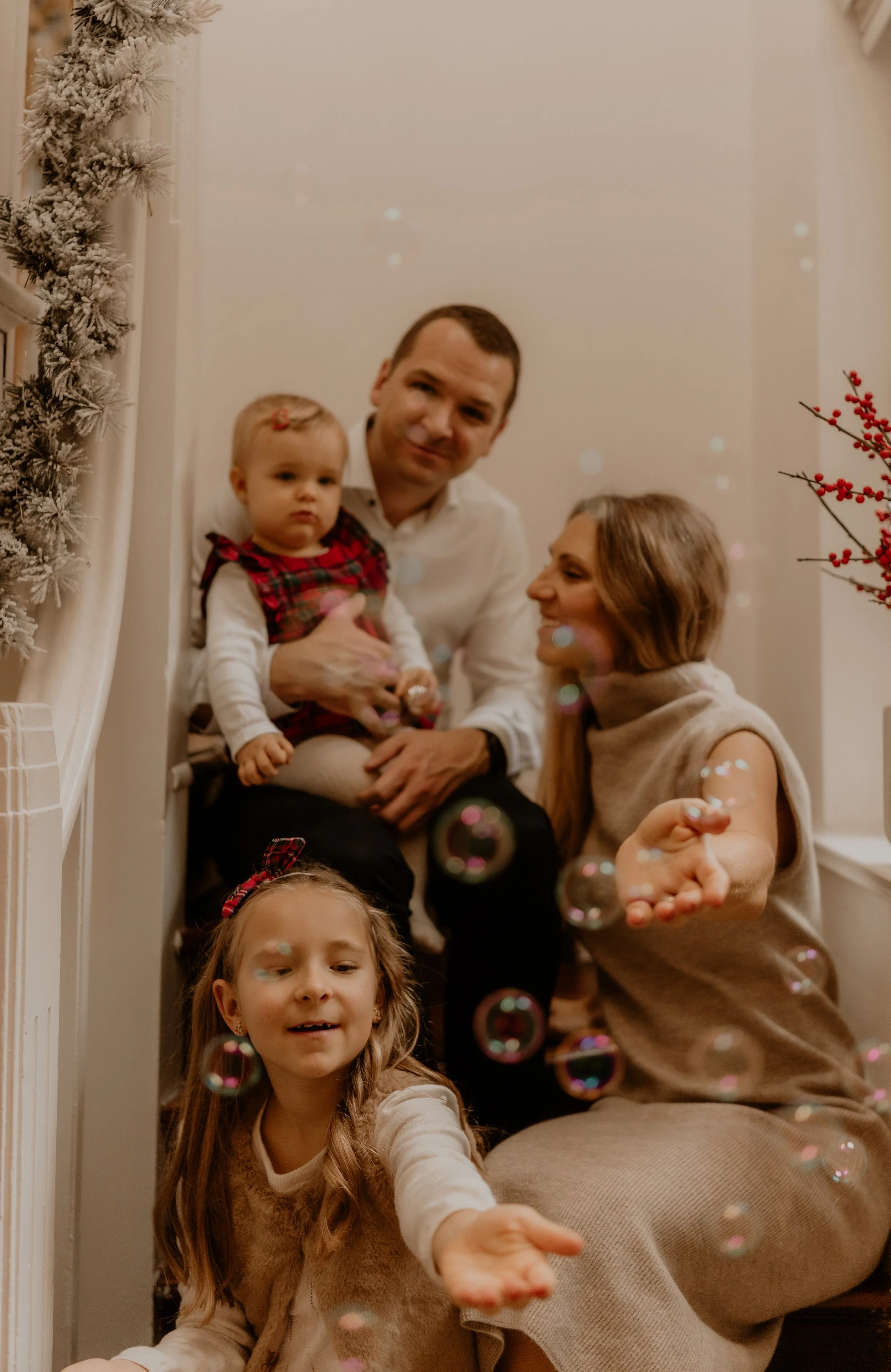 A family of four sitting on stairs decorated for Christmas, with snow-flocked garland and red berries, playing with soap bubbles.