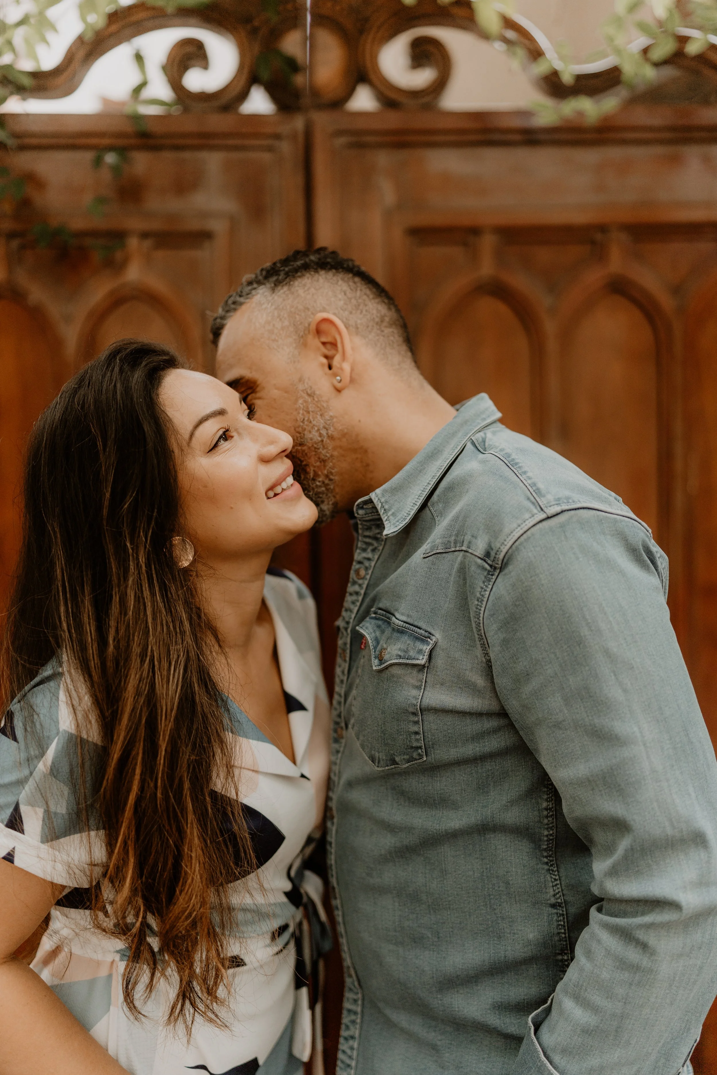 A couple is standing close to each other indoors, smiling and sharing a kiss on the forehead in front of a wooden decorative background