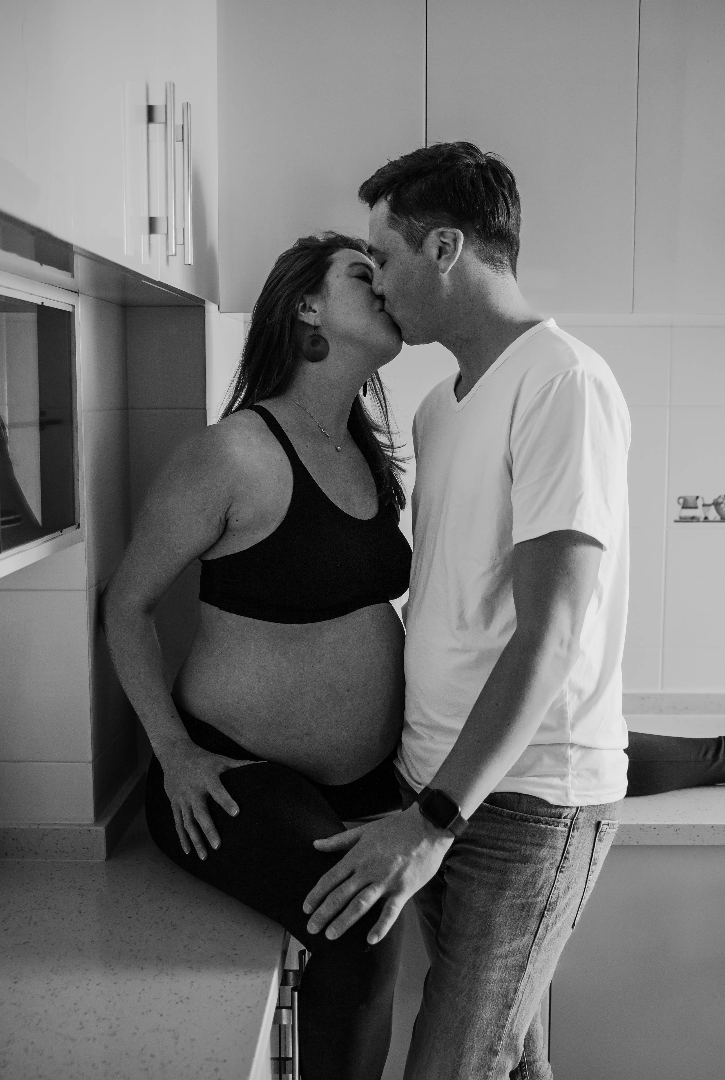 A pregnant woman and a man kiss in a kitchen, with the woman sitting on a countertop and the man standing in front of her, holding her stomach.