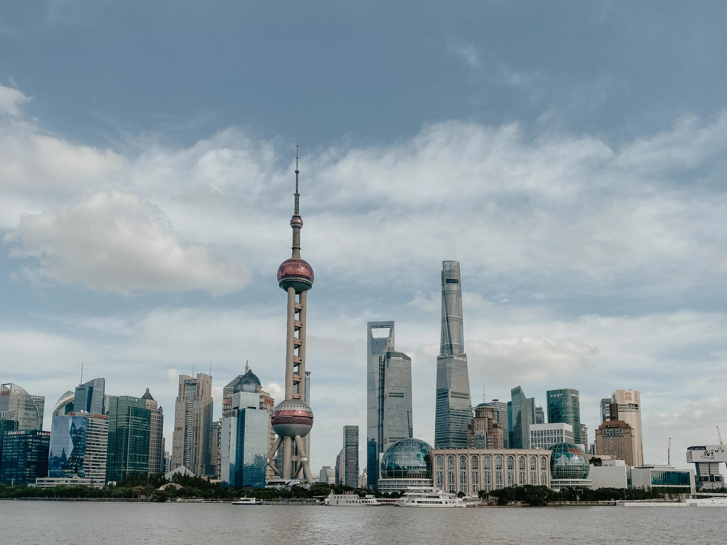 Shanghai skyline featuring the Oriental Pearl Tower, Shanghai World Financial Center, and Shanghai Tower along the Huangpu River under a partly cloudy sky.