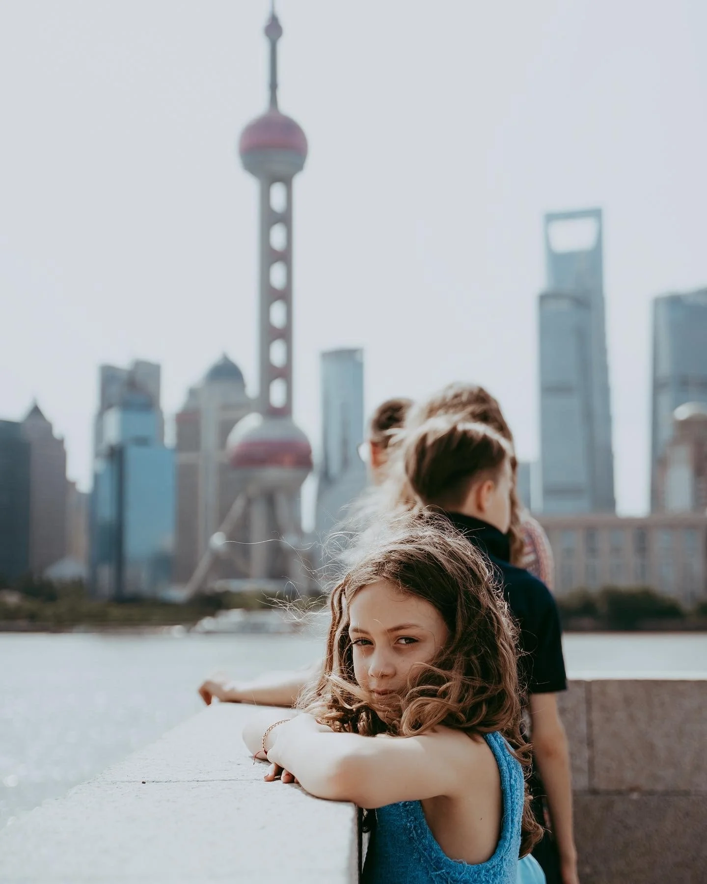 A young girl with curly hair leaning on a stone wall, looking at the camera with a city skyline and the Shanghai Tower in the background.