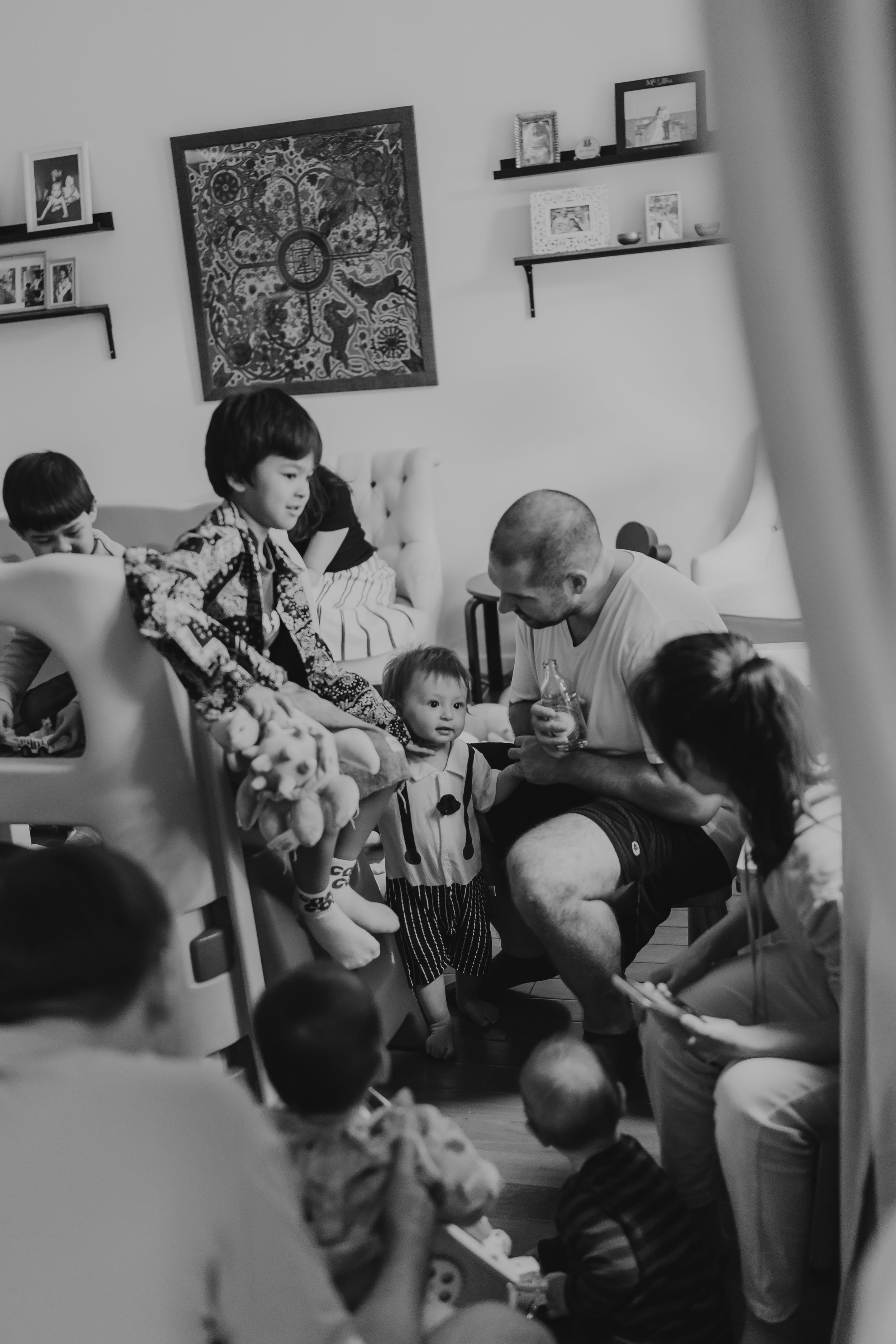 Family gathering in living room with children and adults, some sitting and some standing, engaging with each other. Walls decorated with framed photos and artwork.