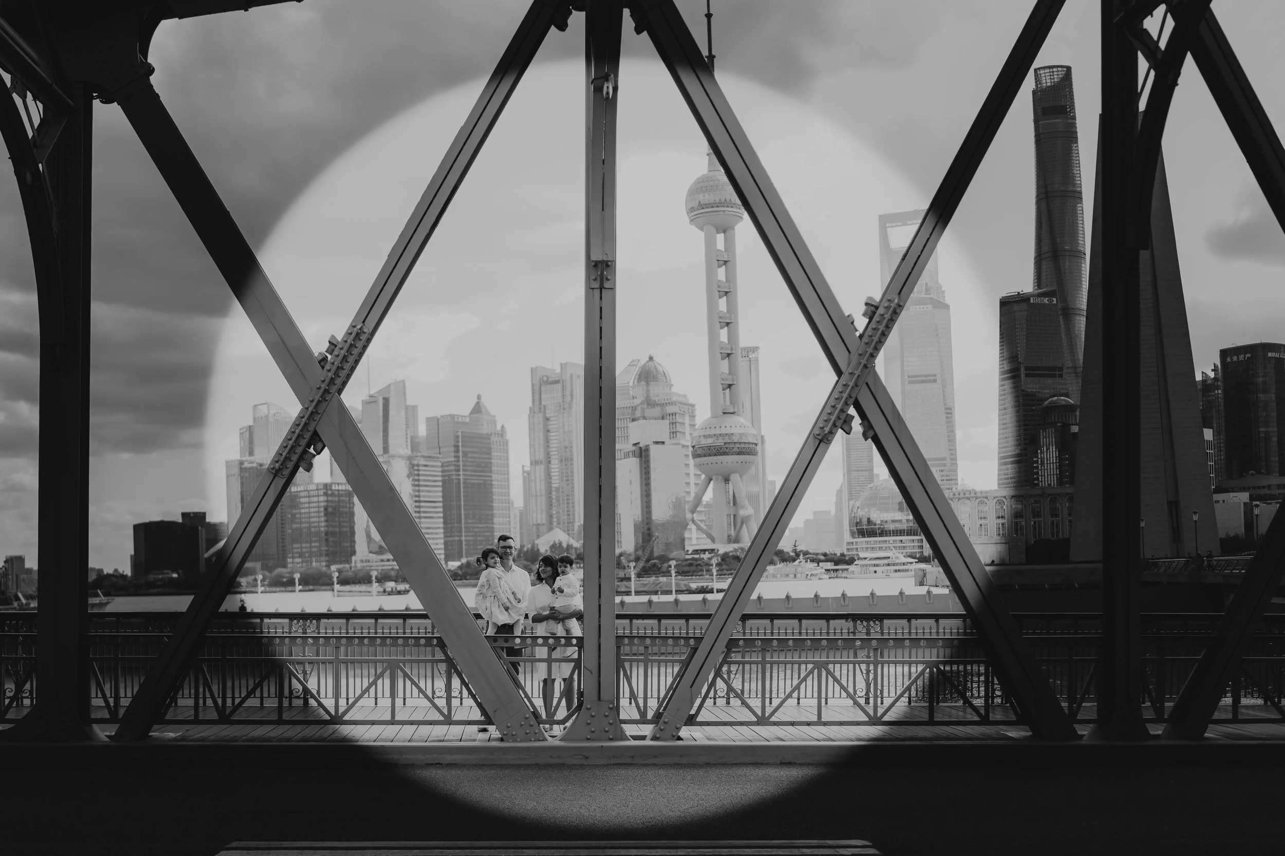 Black and white photo of a city skyline viewed through the steel framework of a bridge, with a group of four people standing on the pedestrian walkway in the foreground.