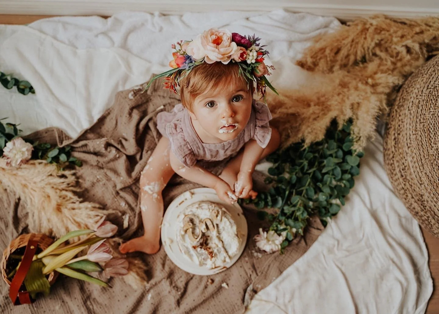 A young child with a flower crown and messy face and hands, sitting on bedding with a cake and flowers around.