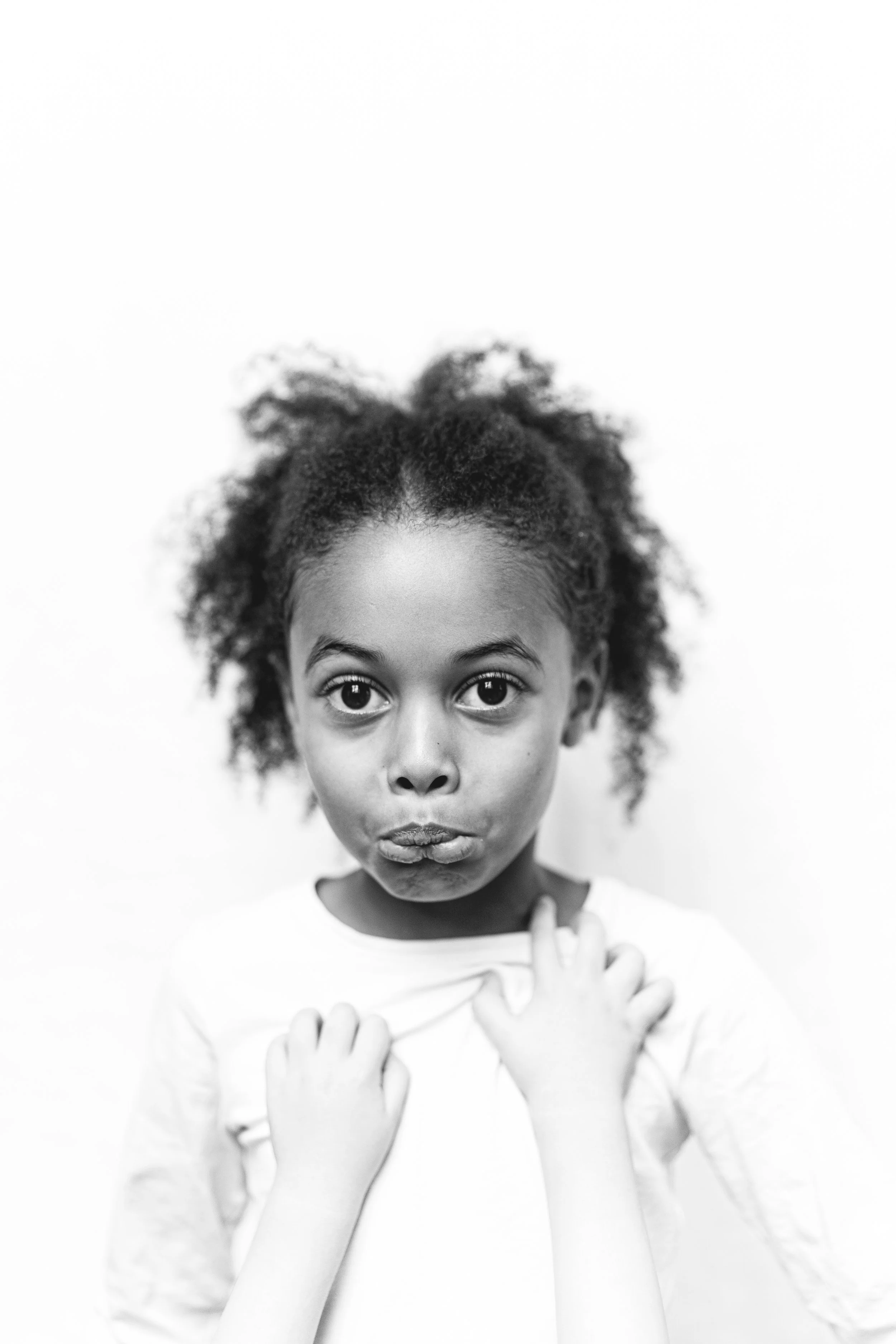 A black-and-white photo of a young girl with curly hair, wide eyes, and a serious expression, holding her shirt with both hands against a plain background. black and white portrait. 