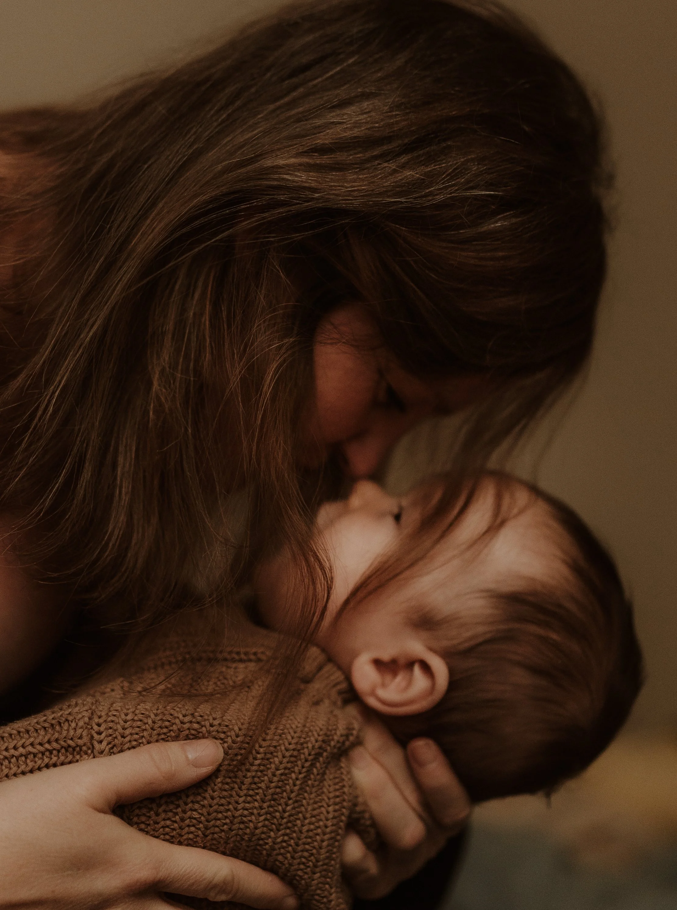 A woman with long brown hair gently holding and kissing a newborn baby dressed in a brown knitted outfit.
