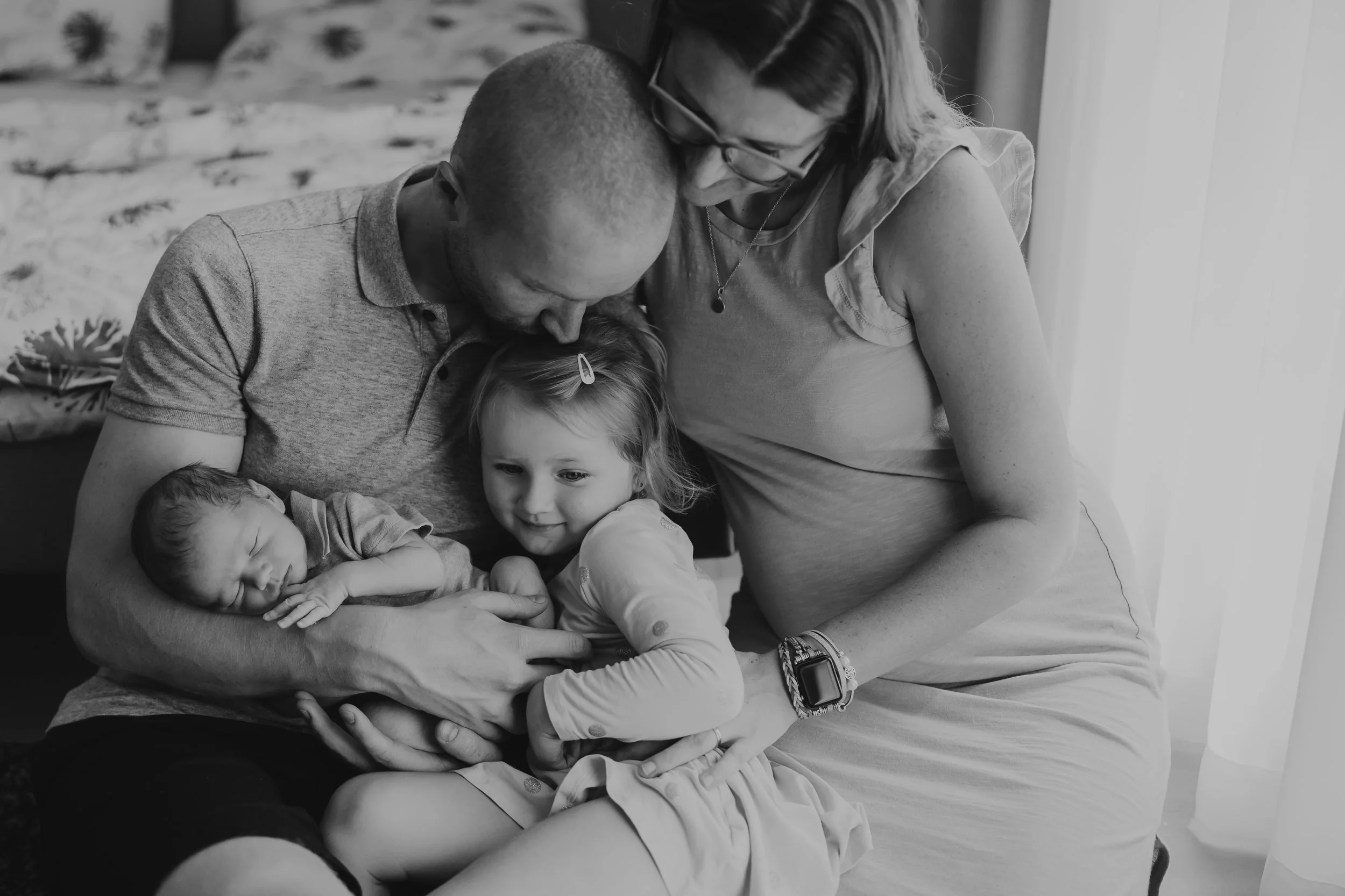 A family of four, including a newborn, is gathered together indoors. The father, mother, daughter, and newborn are closely snuggled, with the father holding the newborn, and the mother and daughter sharing a moment of affection.