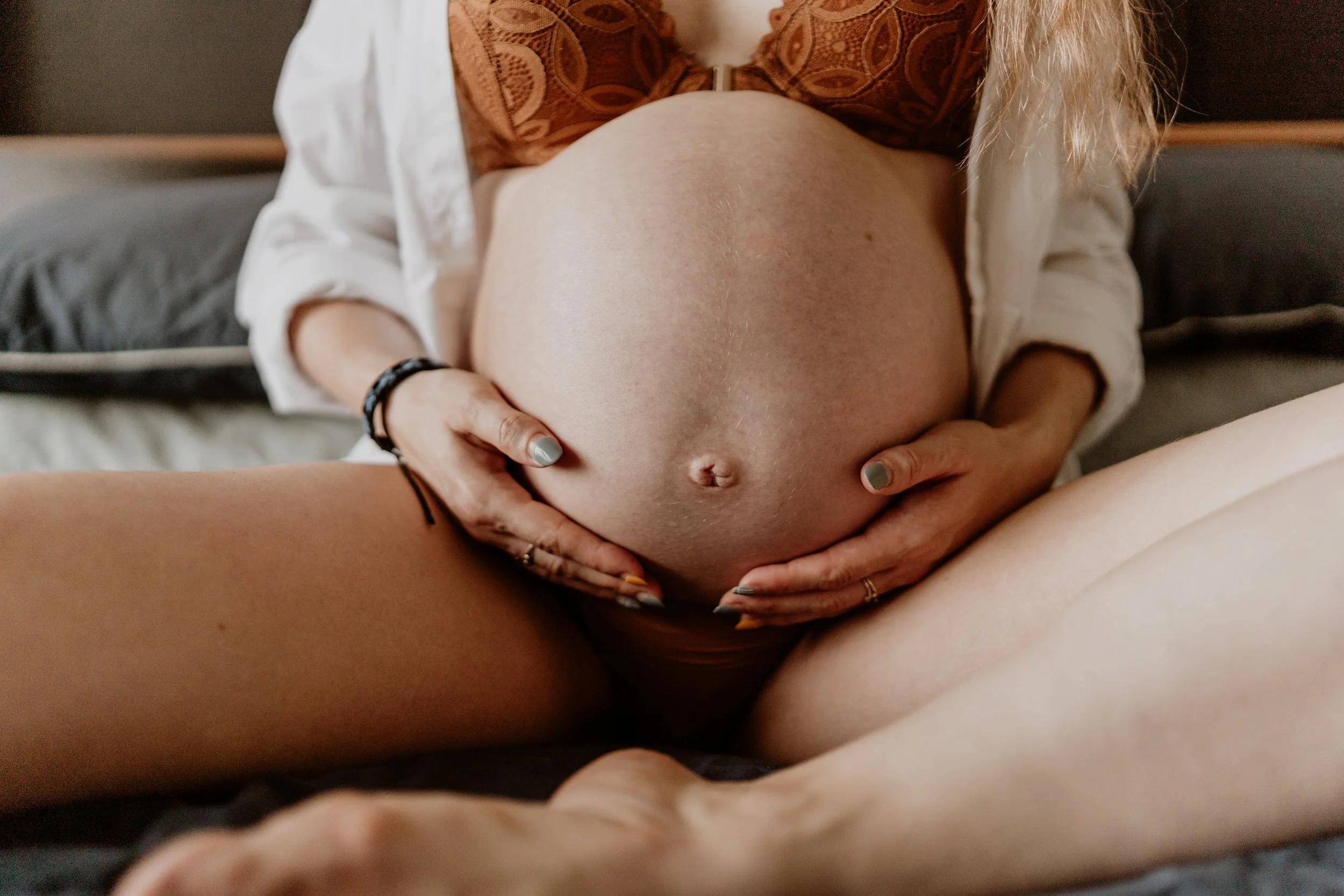 A pregnant woman with her hands on her belly, sitting on a bed with dark pillows in the background.