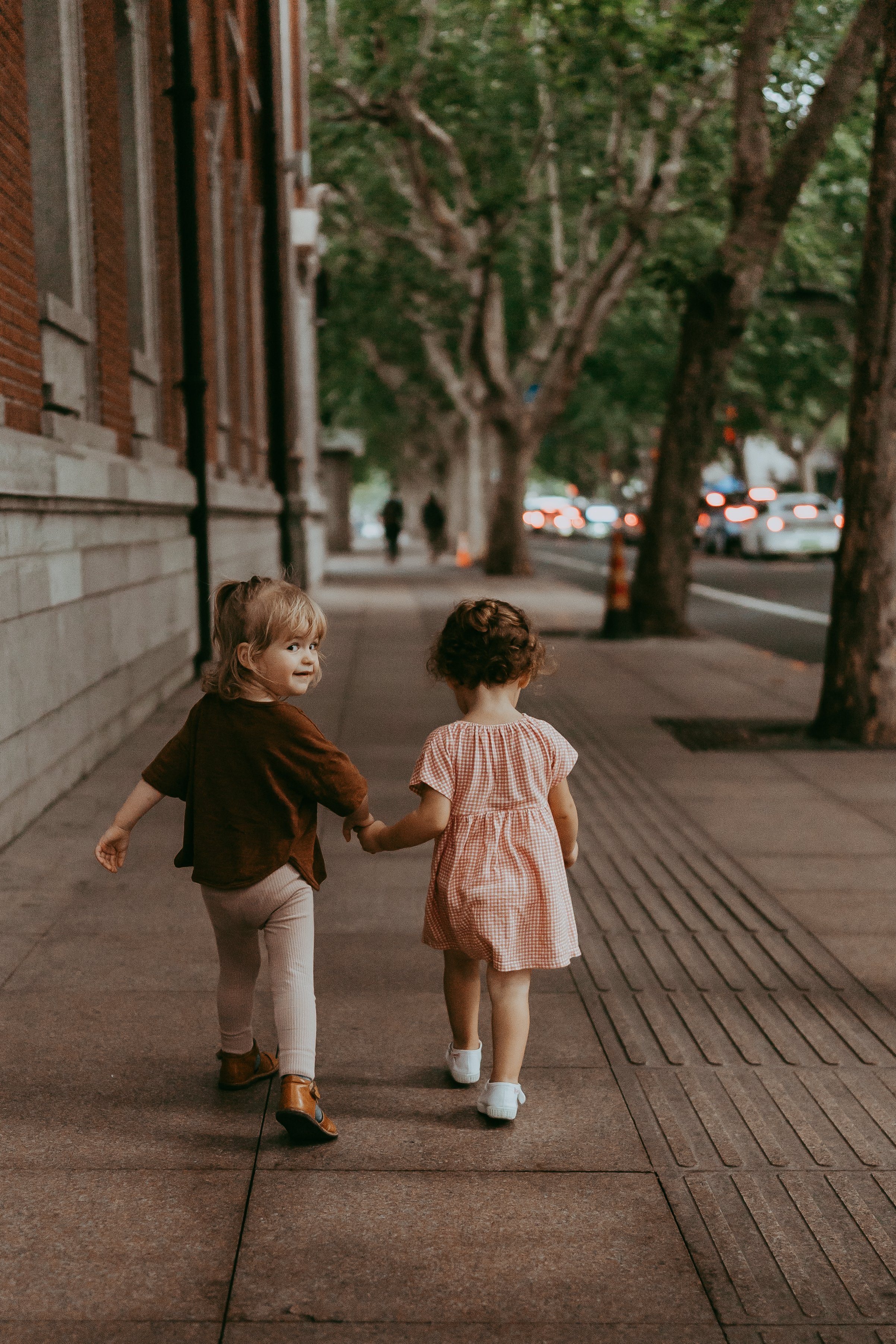 Two young girls walking hand-in-hand on a city sidewalk during dusk, with trees lining the street and blurred cars in the background.