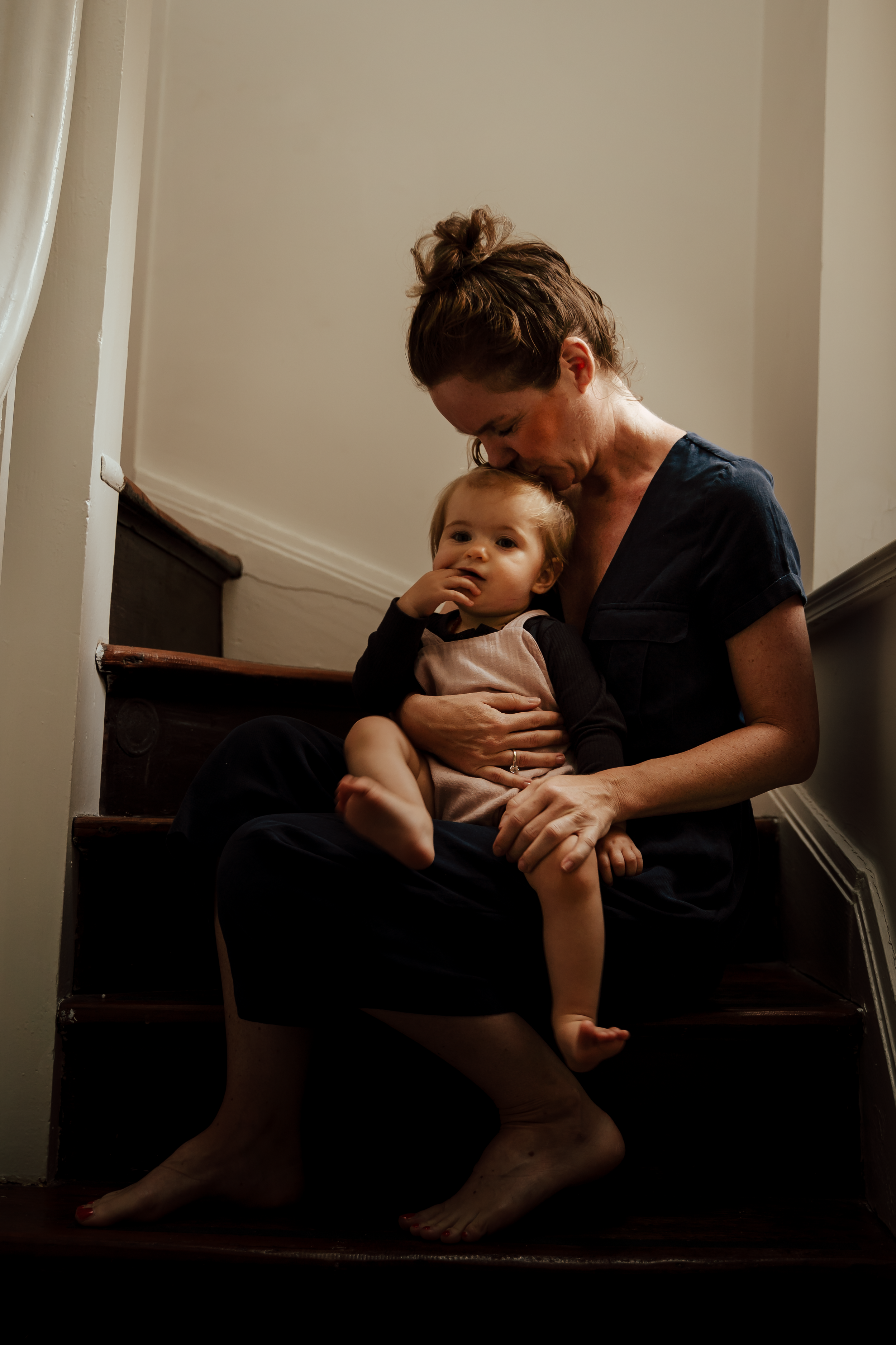 A woman sitting on staircase steps holding a young child with both arms. The woman is kissing the child's forehead while the child has fingers in their mouth. The woman is barefoot, and the setting is indoors with soft lighting.