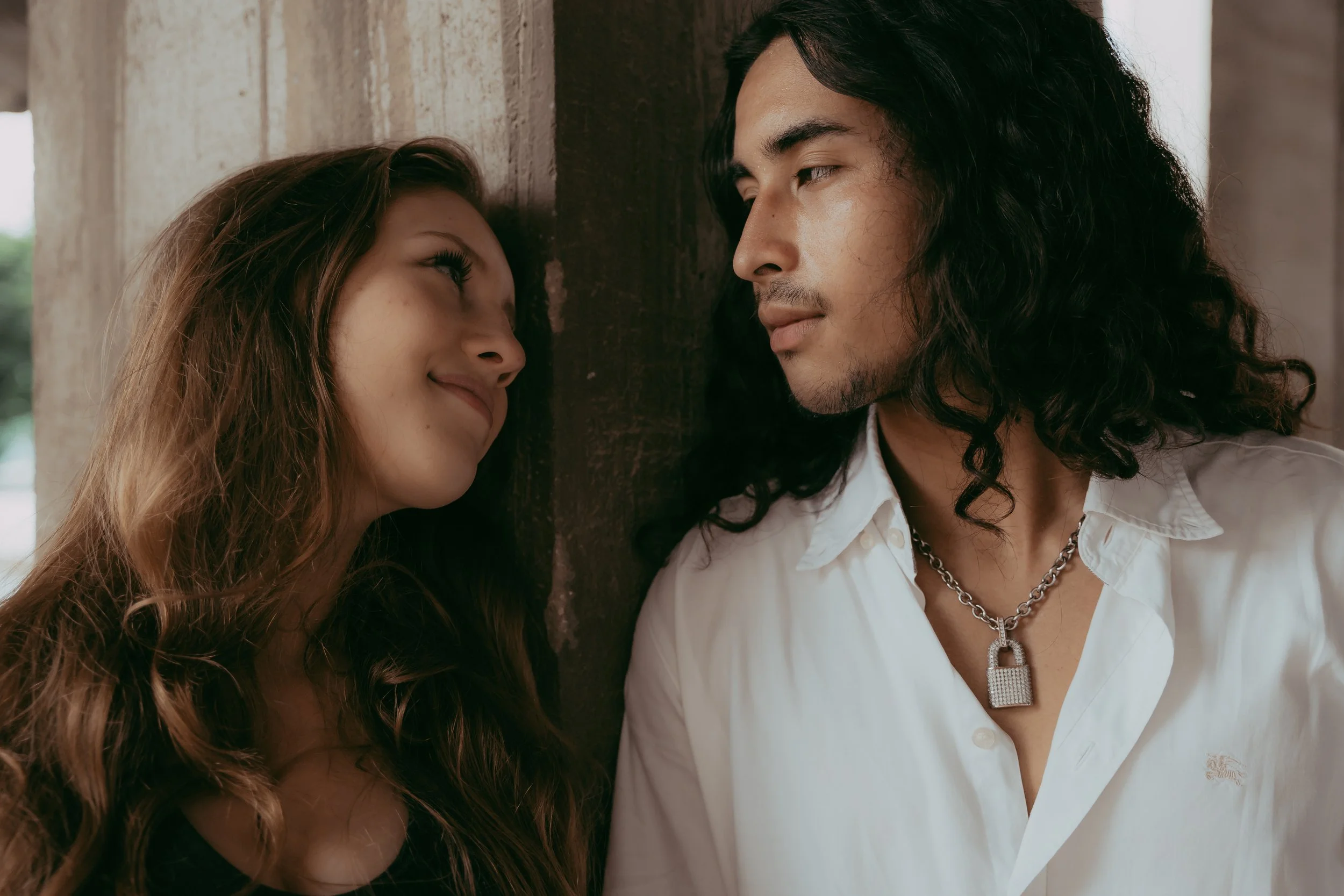 A young woman and a young man with long dark hair leaning against a wall, gazing at each other affectionately.