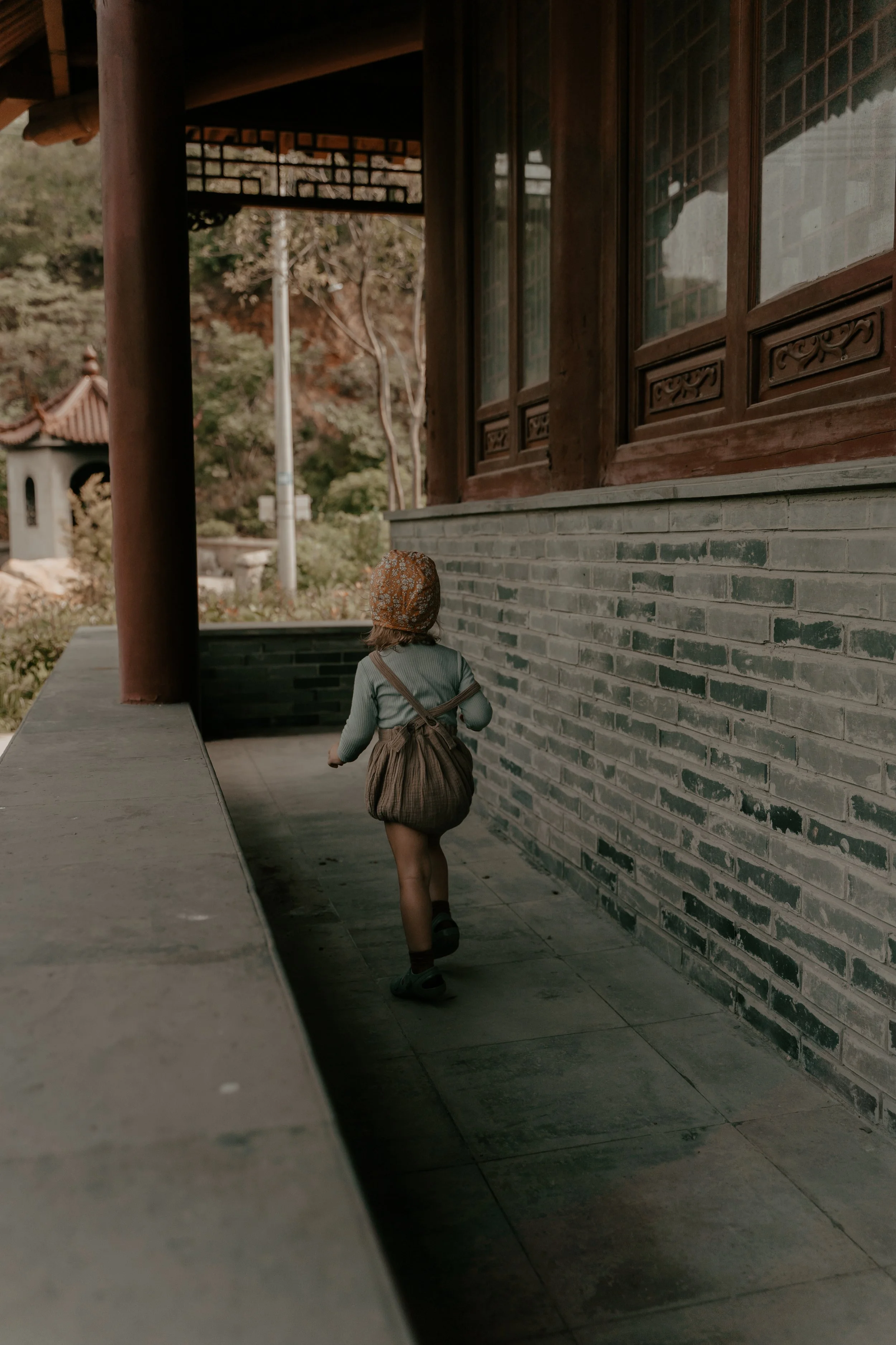 A young child wearing a hat, gray shirt, brown skirt, and carrying a backpack, walking along a covered outdoor corridor of a traditional Asian-style building.