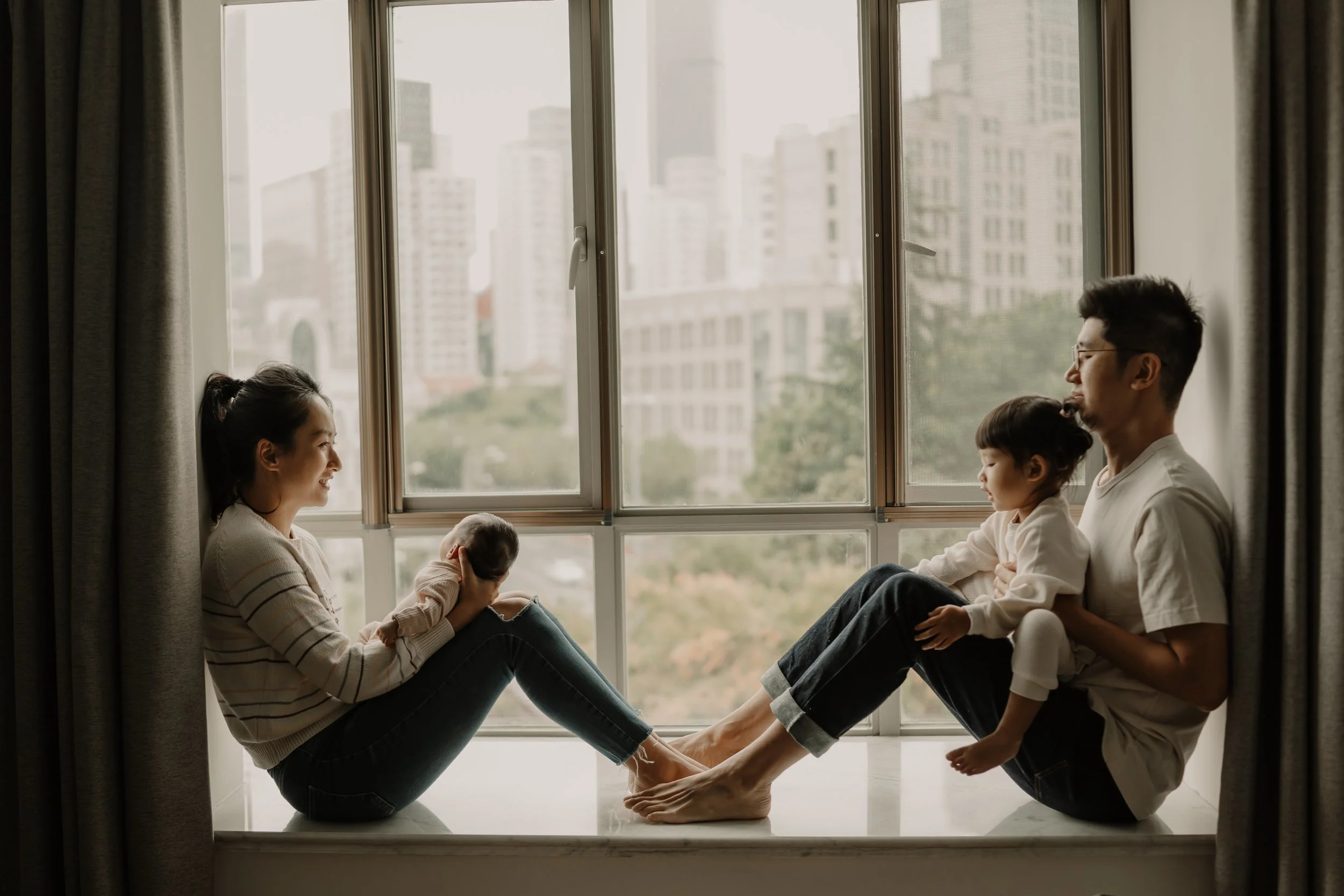 Family sitting on a windowsill, facing each other and holding their babies, with a cityscape view in the background.