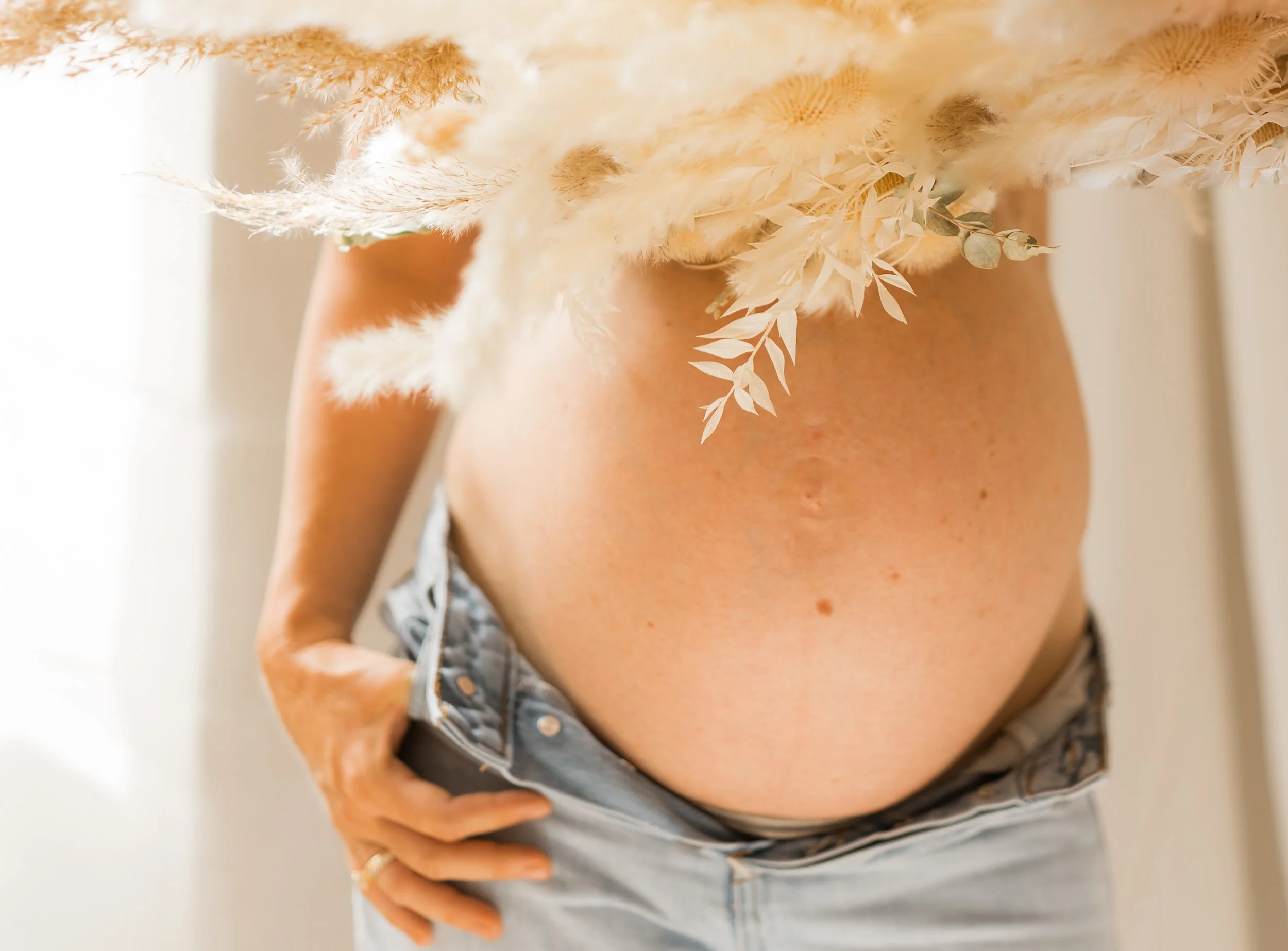 Close-up of a pregnant woman's belly with a floral arrangement covering the top, wearing light-colored jeans.