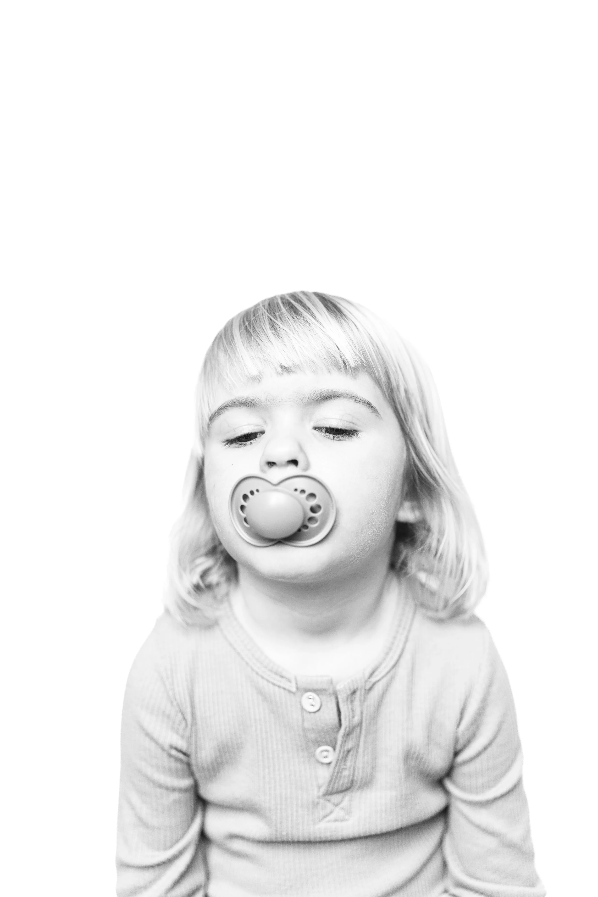 A young child with a pacifier in their mouth, looking downward, wearing a button-up shirt, and has straight, shoulder-length hair. black and white portrait. 
