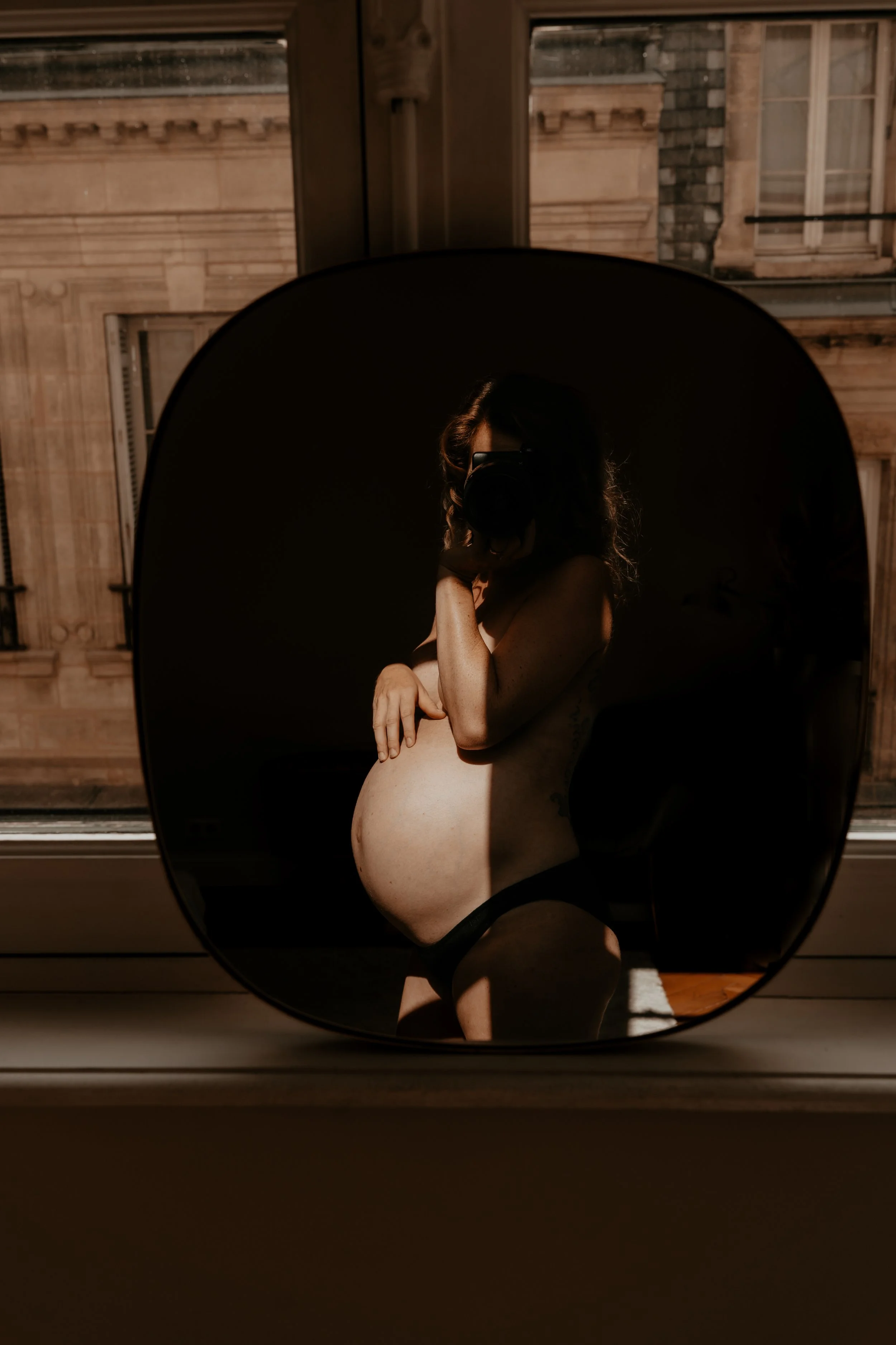 A woman taking a mirror selfie, sitting near a window with sunlight casting shadows, her face partially covered by the camera, with a cityscape visible outside.