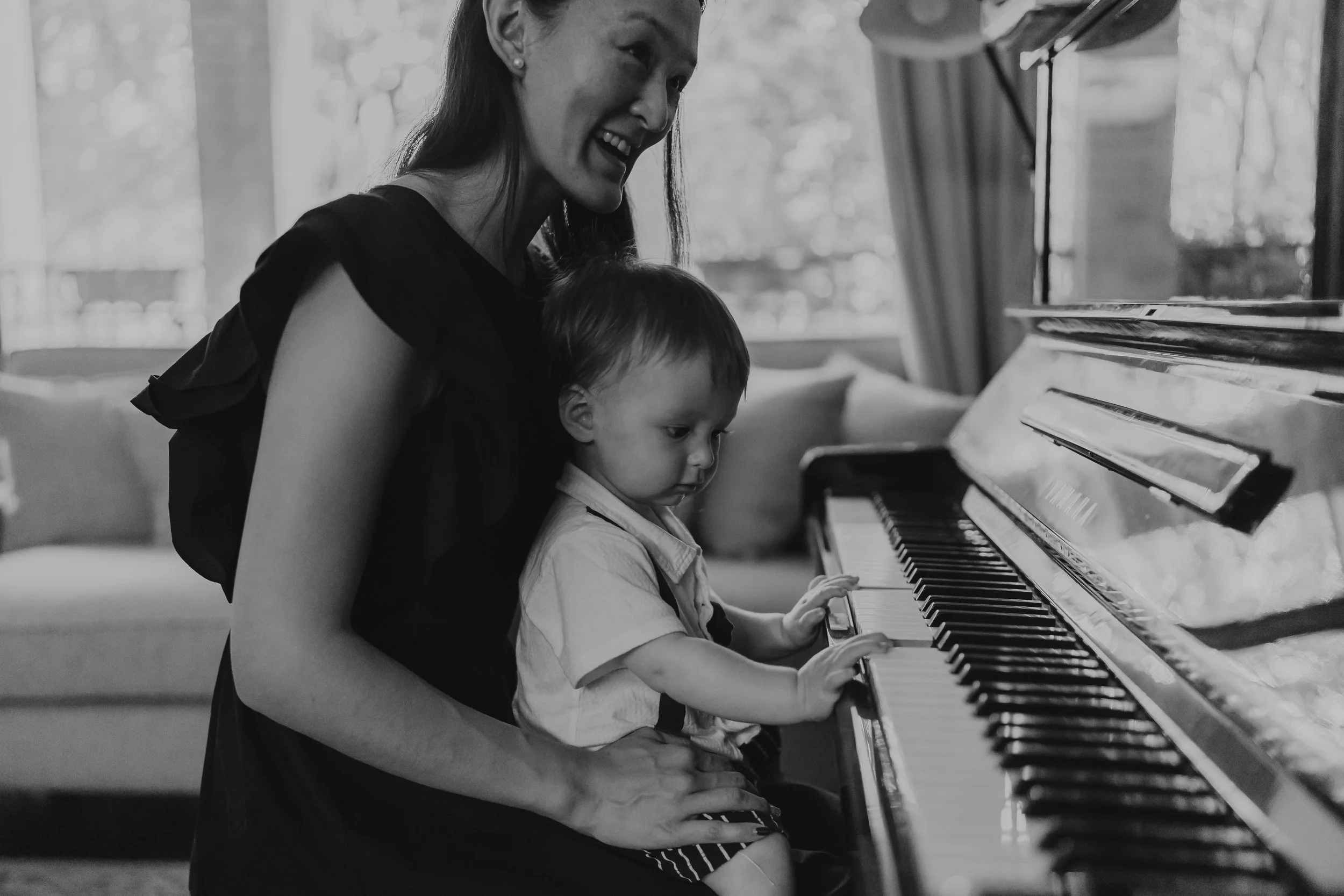 A woman and a young child sitting on her lap playing the piano together in a living room.