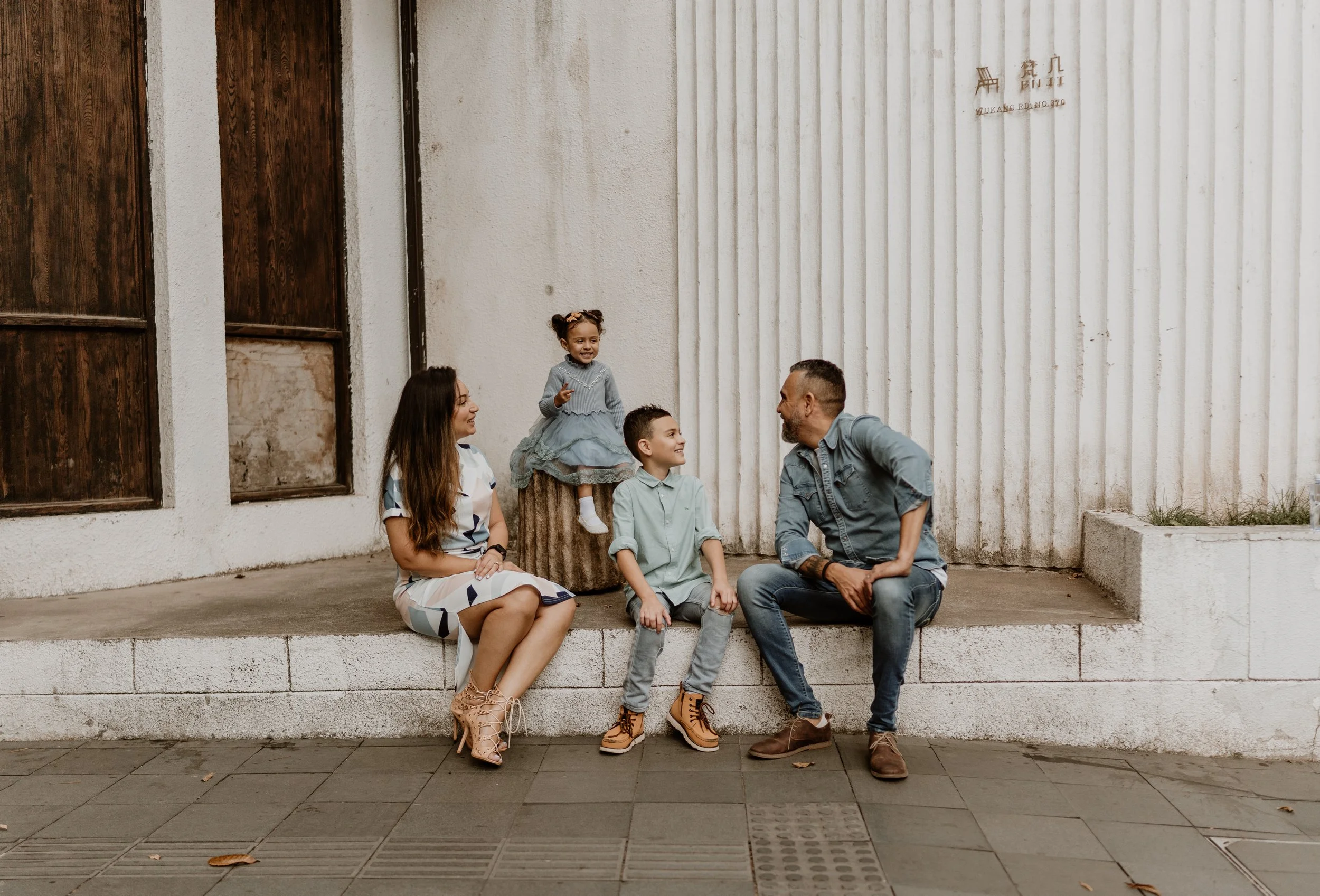 A family sitting outside on a ledge and log, engaging in conversation. The family includes a woman, a man, and two children, one girl and one boy, all smiling and interacting with each other.