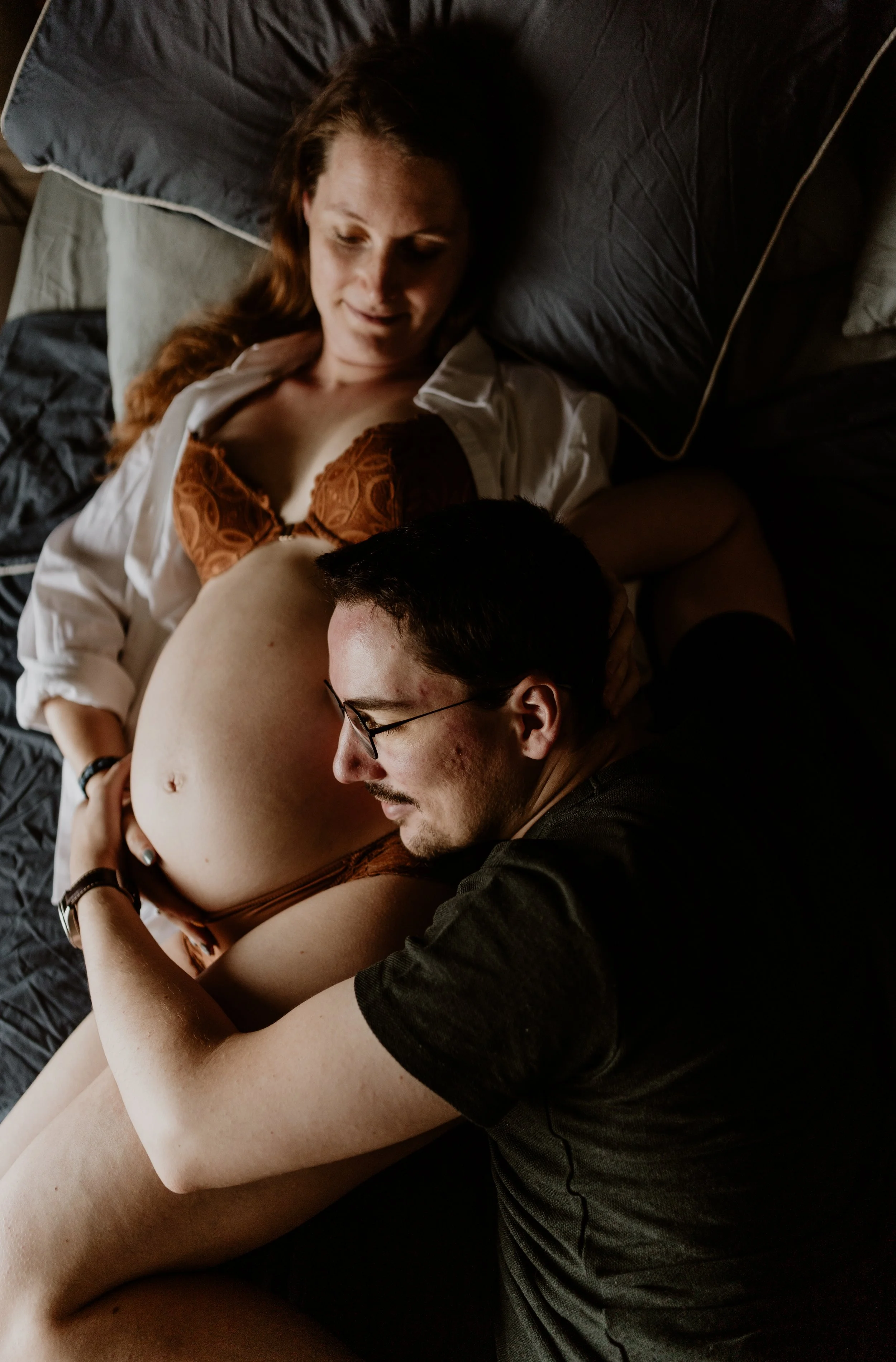 A man and woman lie on a bed together, with the woman appearing pregnant and the man resting his head on her belly.