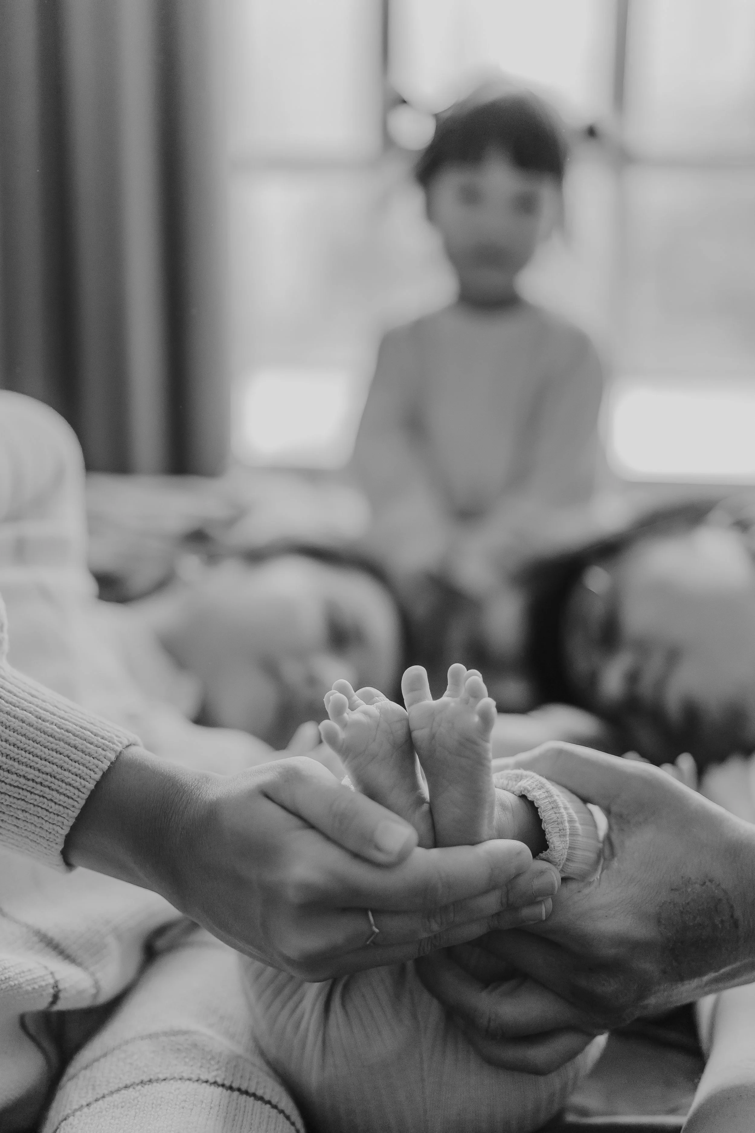 Close-up of a newborn baby's hands being gently held by an adult's hand, with two children lying in bed in the background.