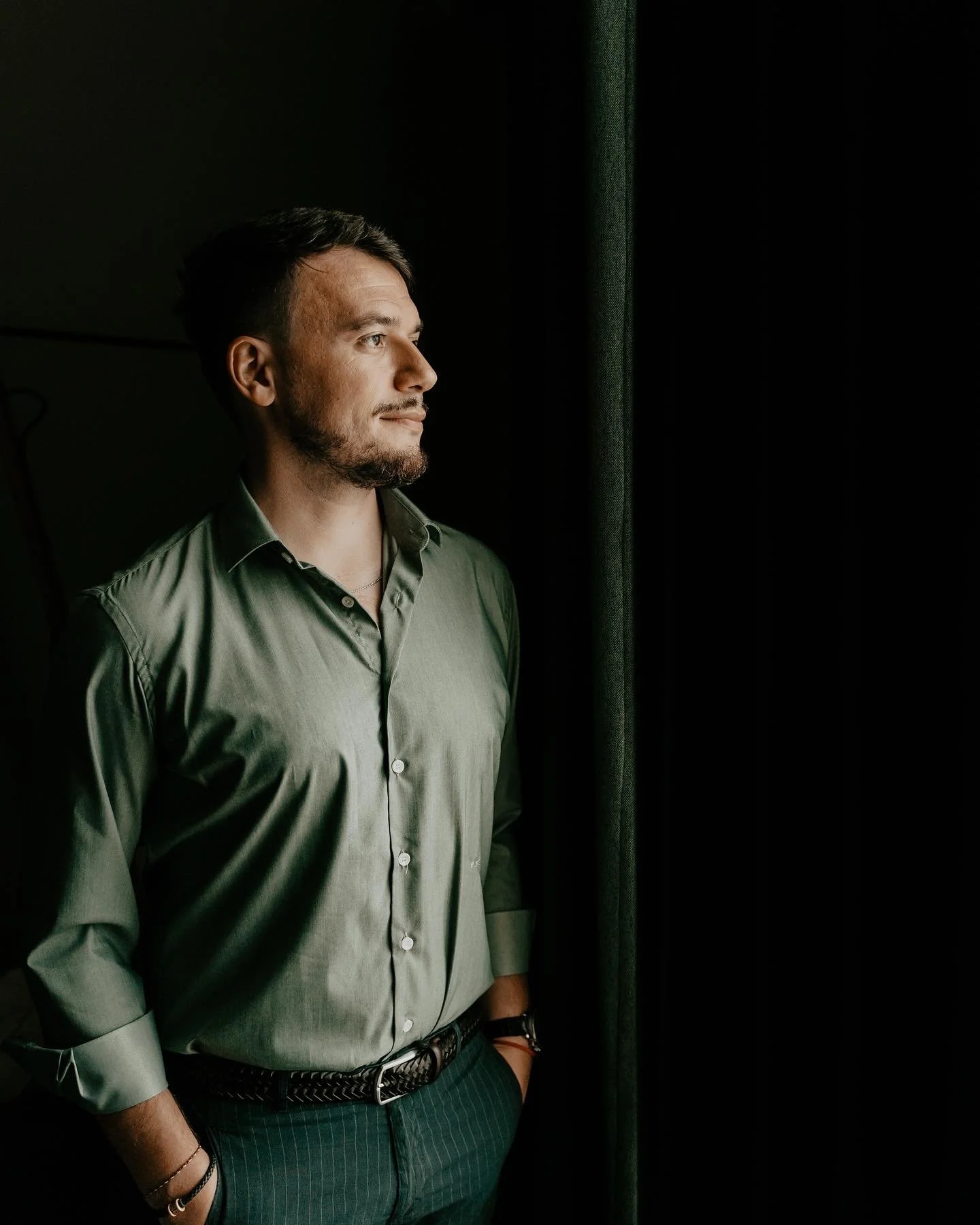 A man standing by a window, looking outside, dressed in a green shirt and dark pants, with his hands in his pockets.