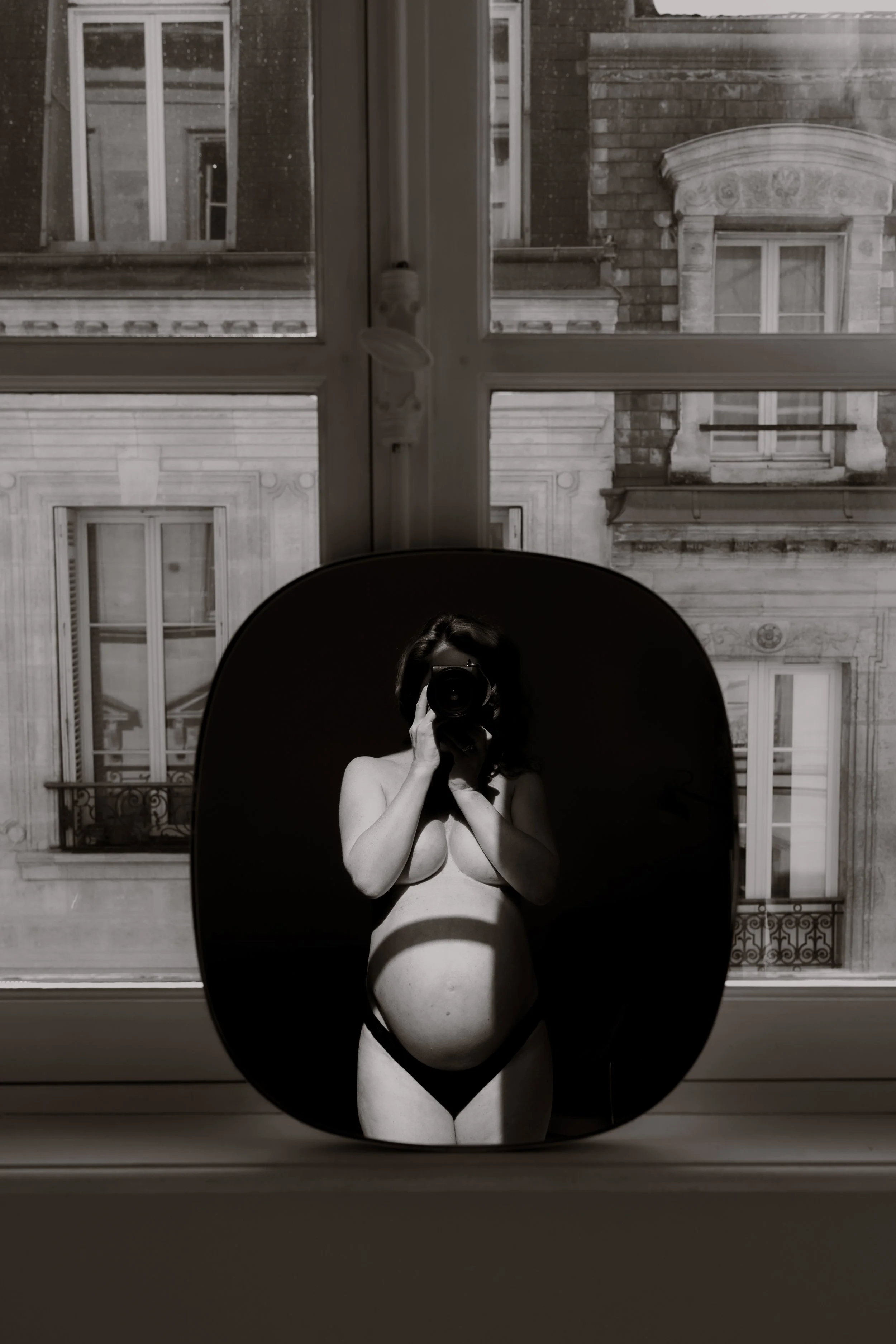 Black and white reflection of a pregnant woman taking a photo of herself with a camera in a round mirror placed on a windowsill, with buildings visible outside the window.