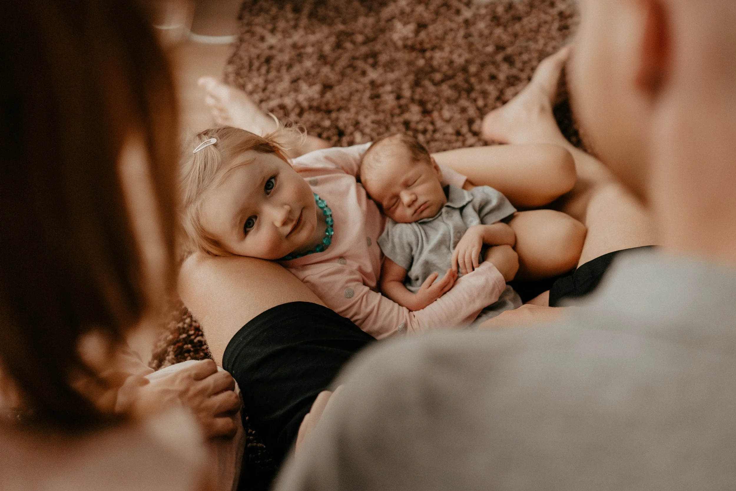 A young girl with blonde hair and a child with light brown hair lying on a person's lap on a carpeted floor, with the girl looking at the camera and the child sleeping peacefully.