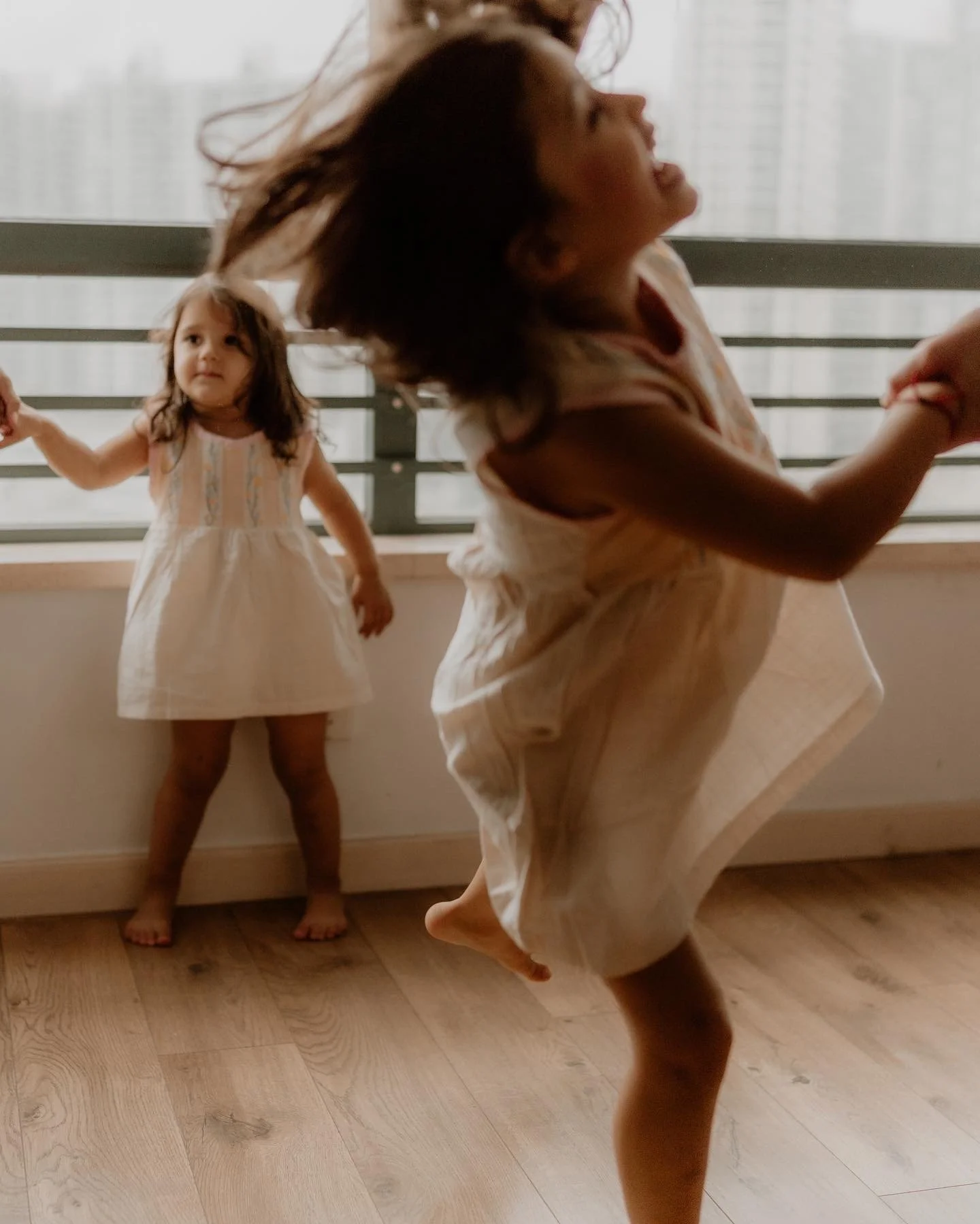 Two young girls in white dresses are playing indoors, holding hands. One girl is in mid-air, jumping happily, while the other girl stands near a window holding a hand and looking on.