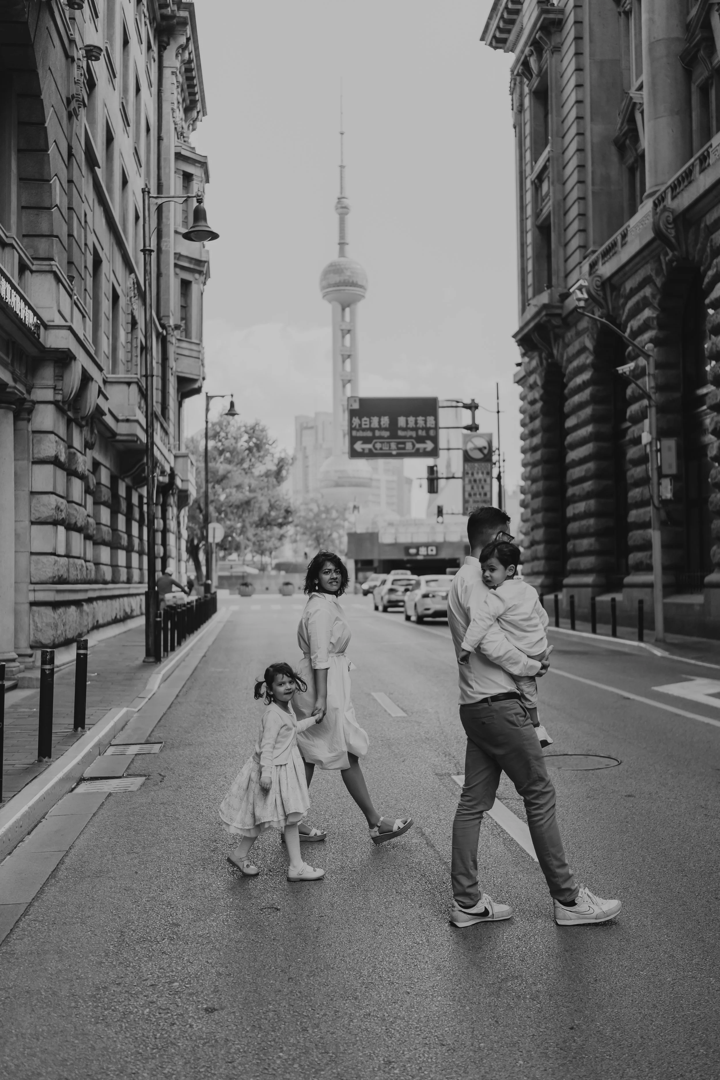 A family crossing a city street with tall buildings and a tower in the background, in black and white.