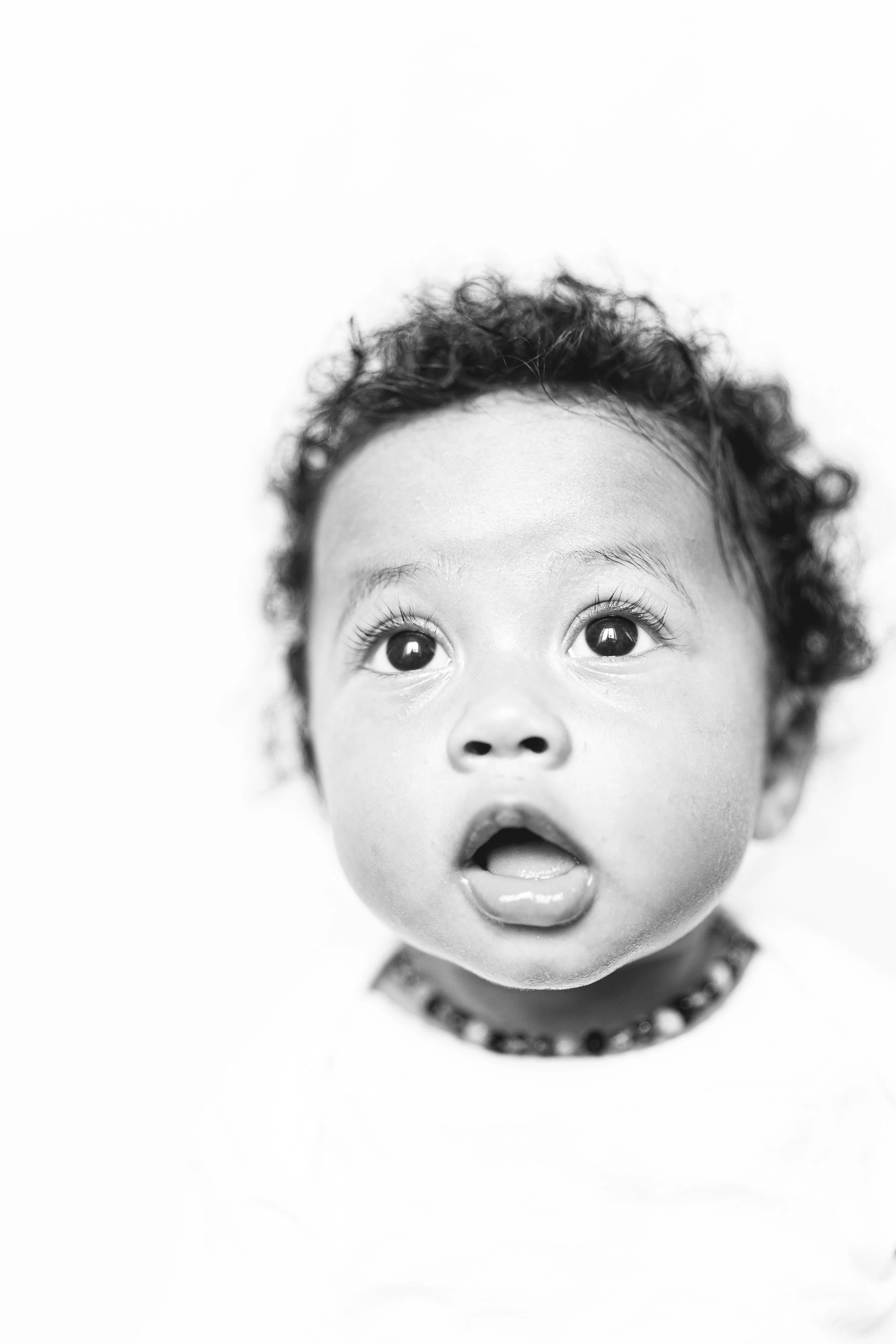 Black and white close-up of a young child's face with wide eyes and mouth slightly open, curly hair, and a necklace visible at the bottom.