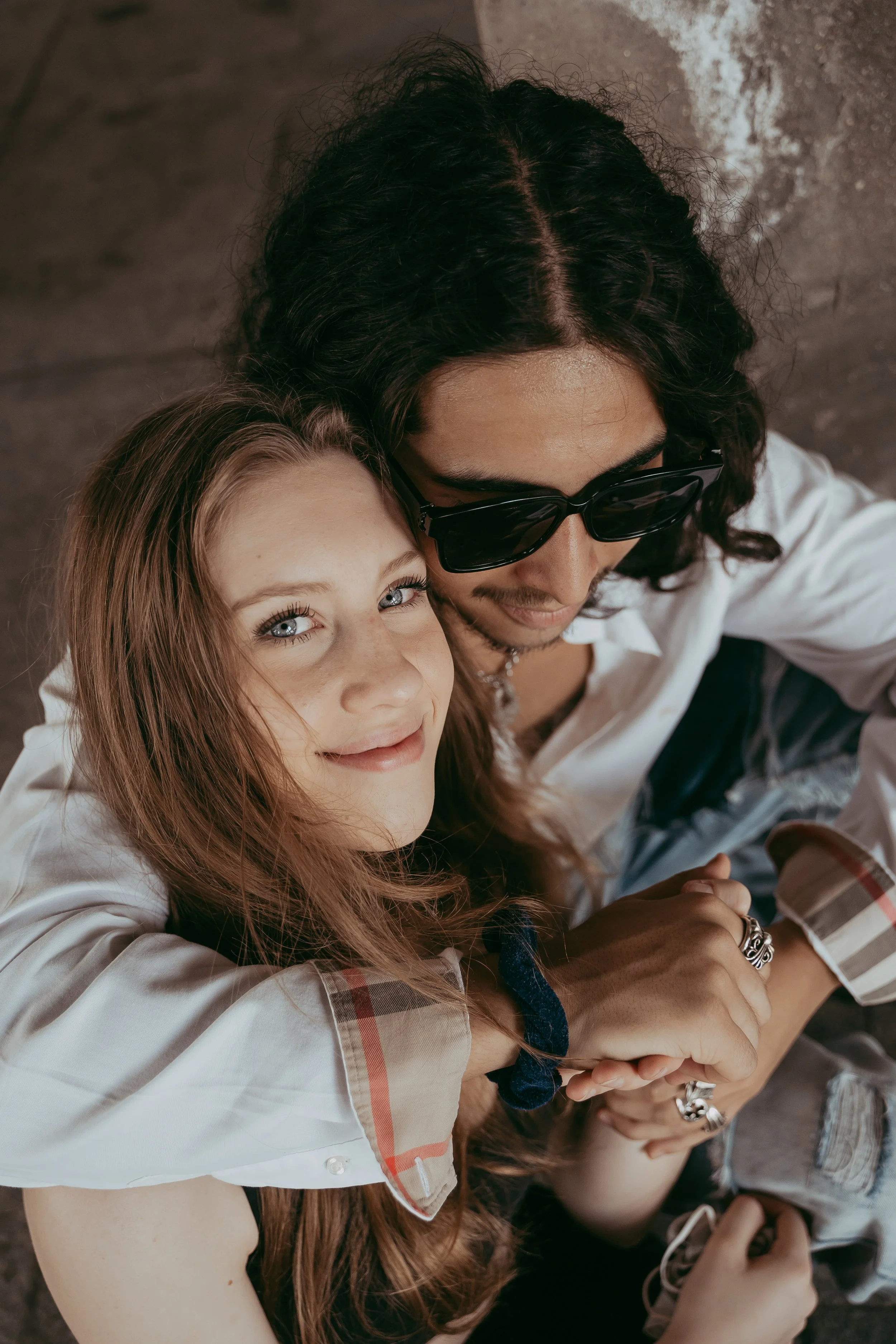 A young woman with reddish hair and blue eyes smiling, hugging a man with long dark hair, wearing sunglasses and a white shirt, sitting close together indoors.