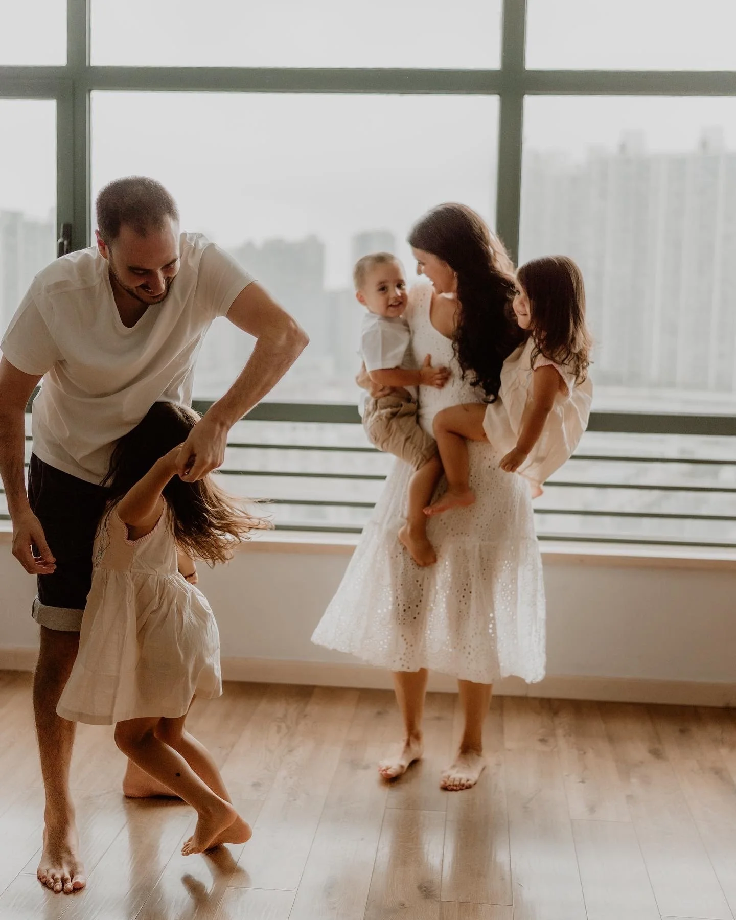 A family of four dancing together indoors near a large window with a cityscape view in the background.