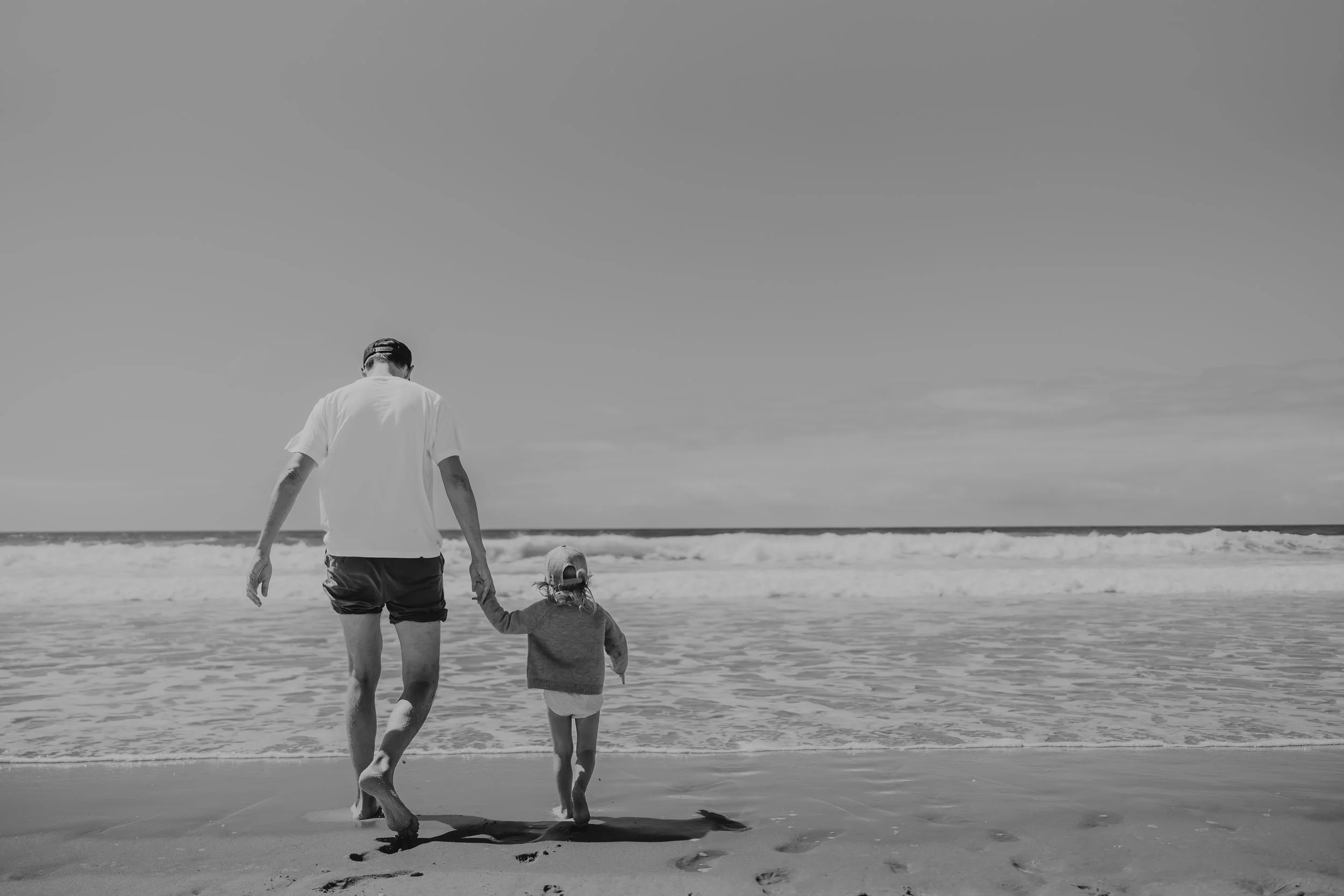 A man and a young girl holding hands walking along the beach with waves crashing in the background.