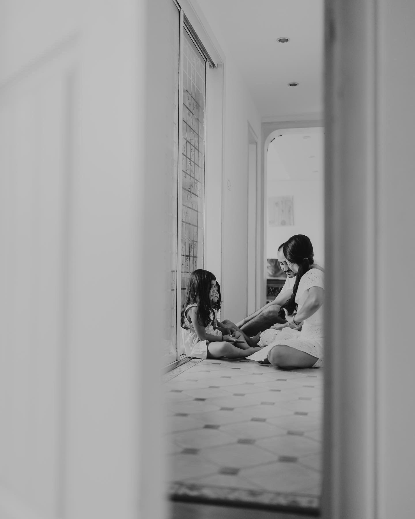 A woman and a young girl sitting on a hallway floor, engaged in conversation, as seen through a doorway, in black and white.