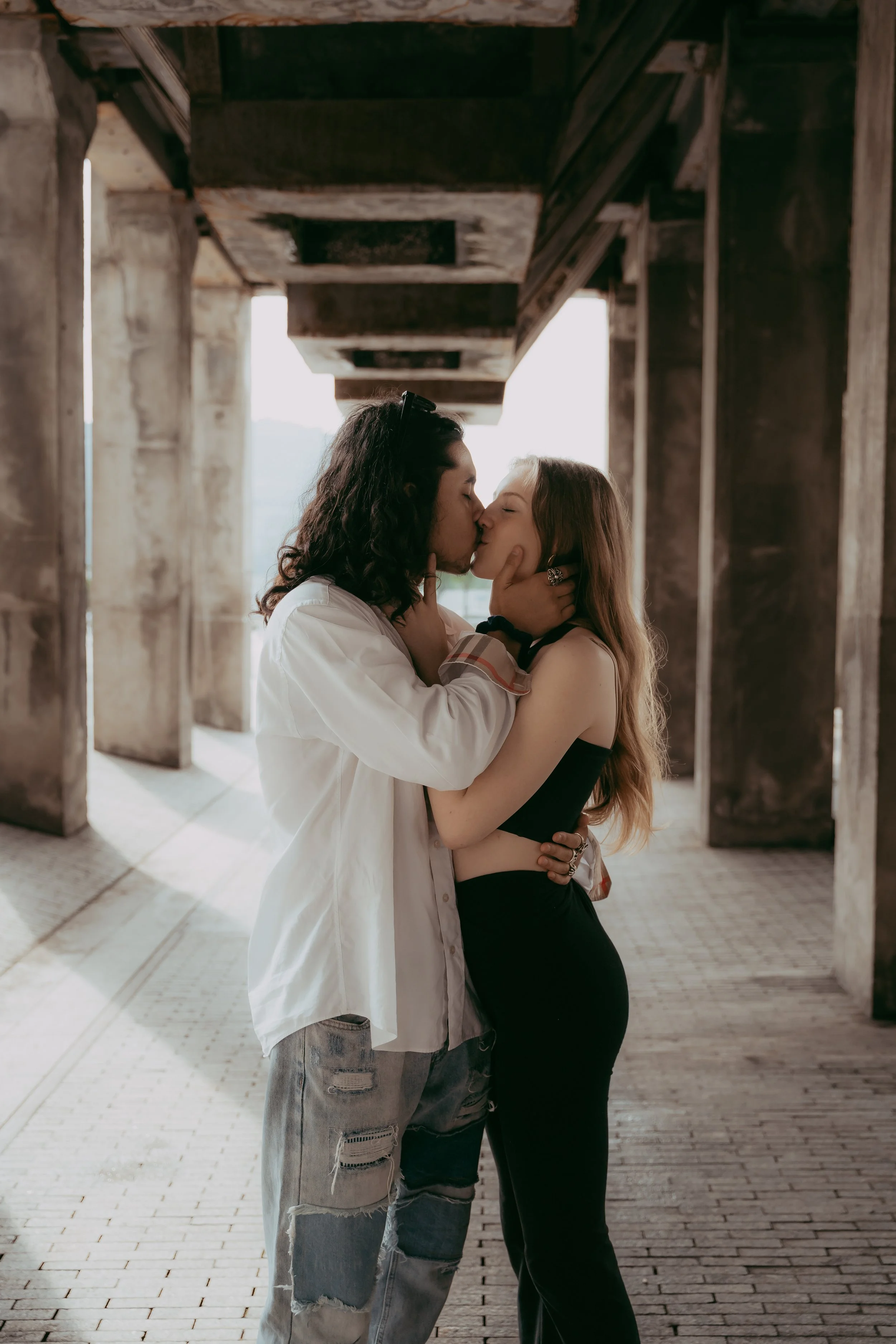 A couple sharing a kiss under a concrete bridge, with the man wearing a white shirt and ripped jeans, and the woman in a black sleeveless top and black pants.