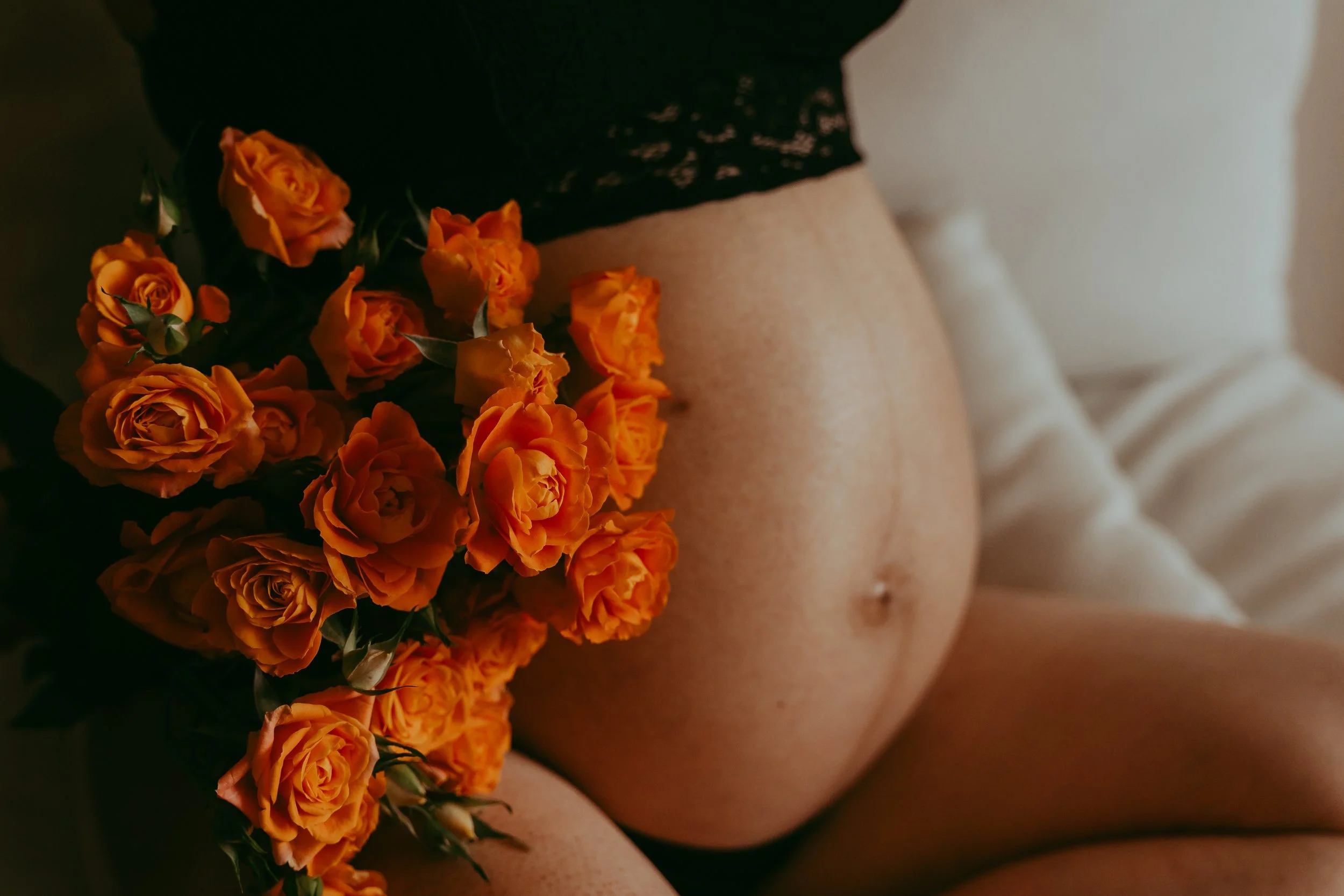 Close-up of a pregnant belly with a bunch of orange roses resting on it, part of a person's torso and thigh visible, black lace clothing at the top, background includes a white bedding surface.