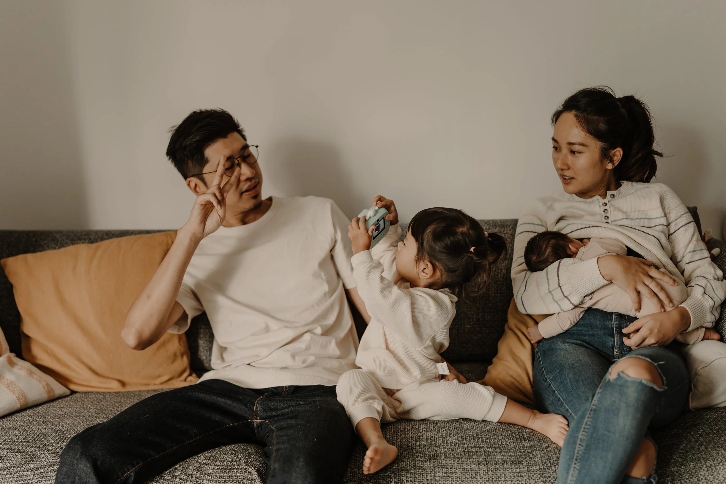 Family of four on a gray couch; father and young daughter playing with camera, mother breastfeeding baby, warm-toned living room