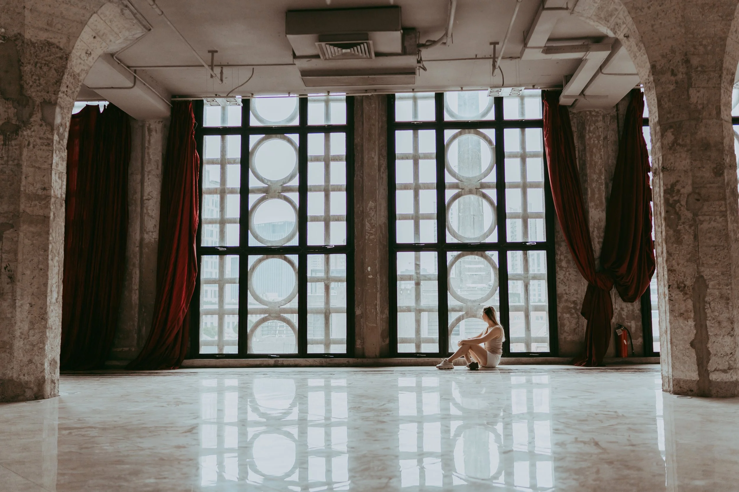 A woman sitting alone on the floor of a large, industrial-style room by a window with a geometric pattern, with red curtains on either side, and reflected on the shiny marble floor.