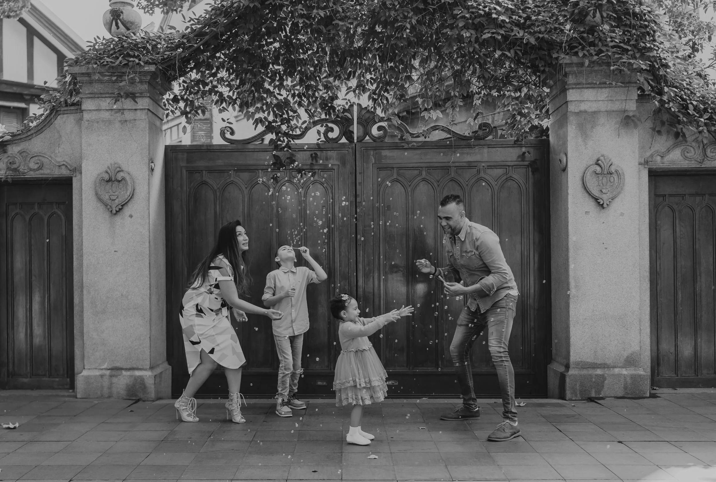 A family of five, including two children, is playing with bubbles in front of a large wooden gate, smiling and laughing.