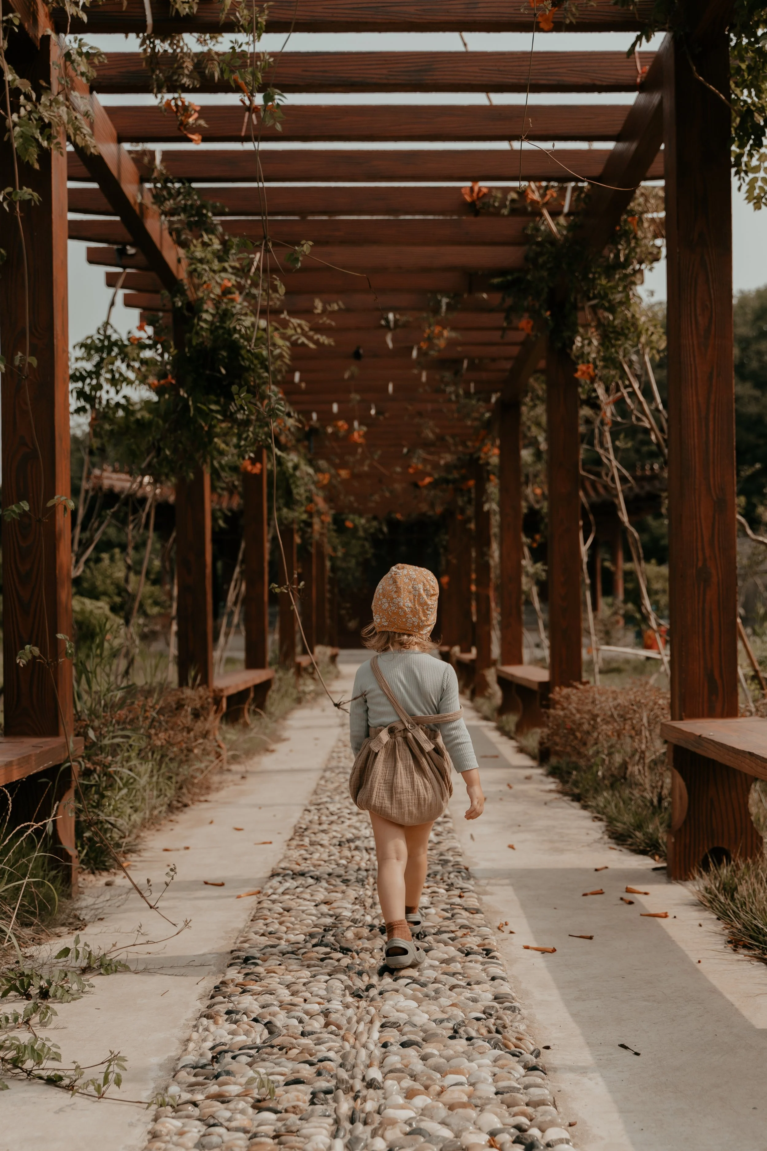 A child walking down a stone and pebble pathway beneath a wooden pergola decorated with plants and hanging lights.