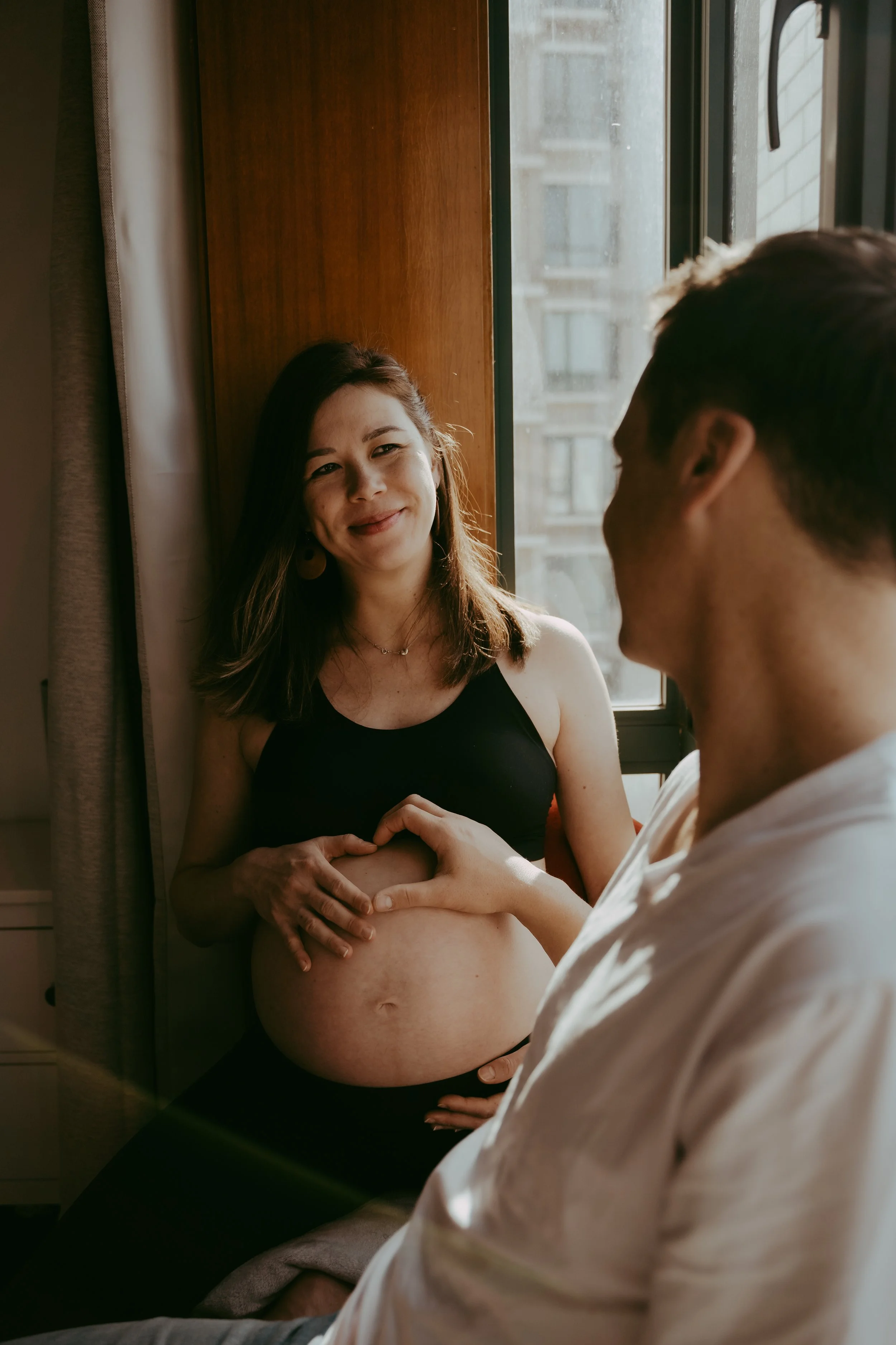 Pregnant woman smiling as her partner gently touches her belly inside a room with sunlight streaming through a window.