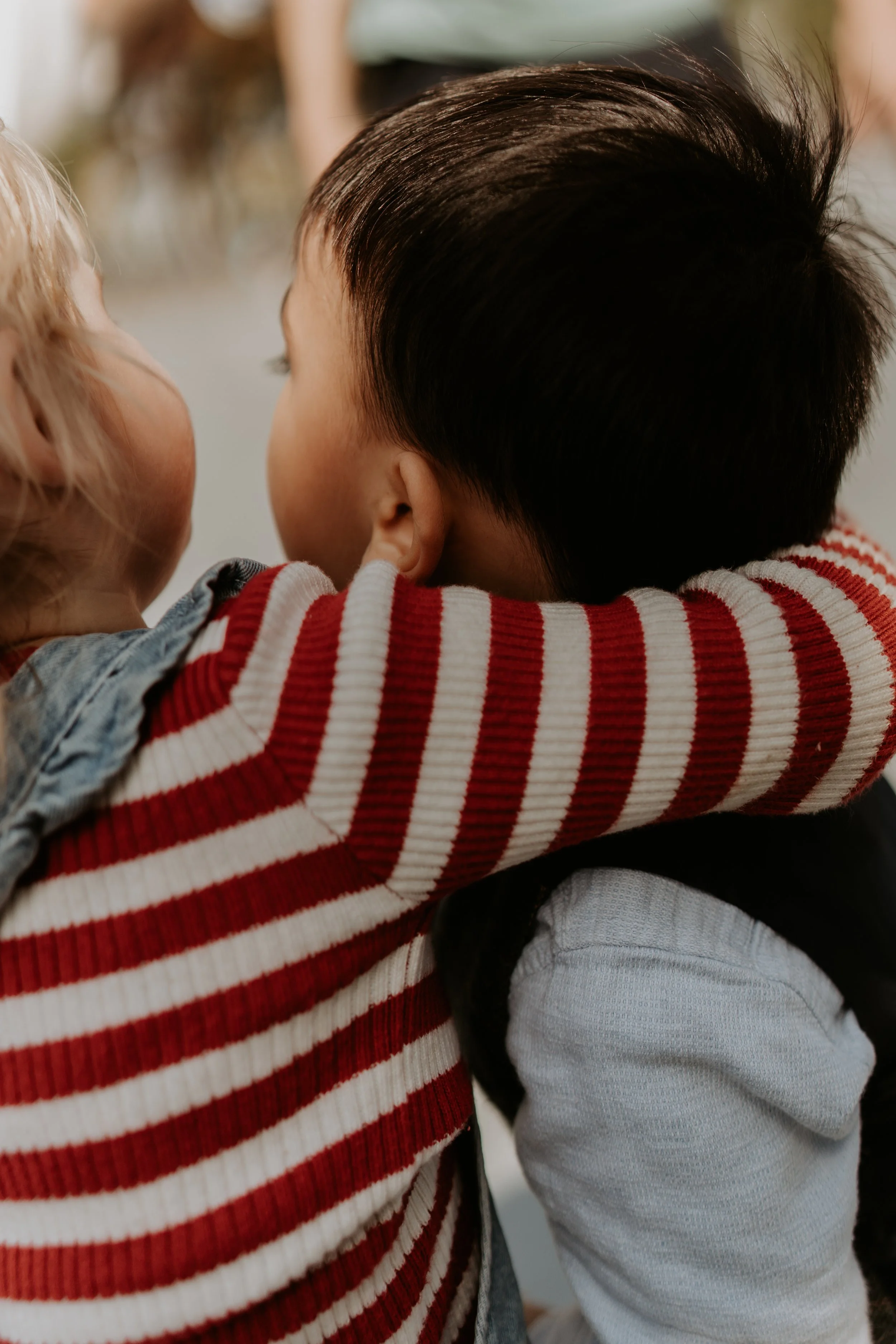 A person with short dark hair and a striped red and white long sleeve hugging a young child with blonde hair and a denim jacket.