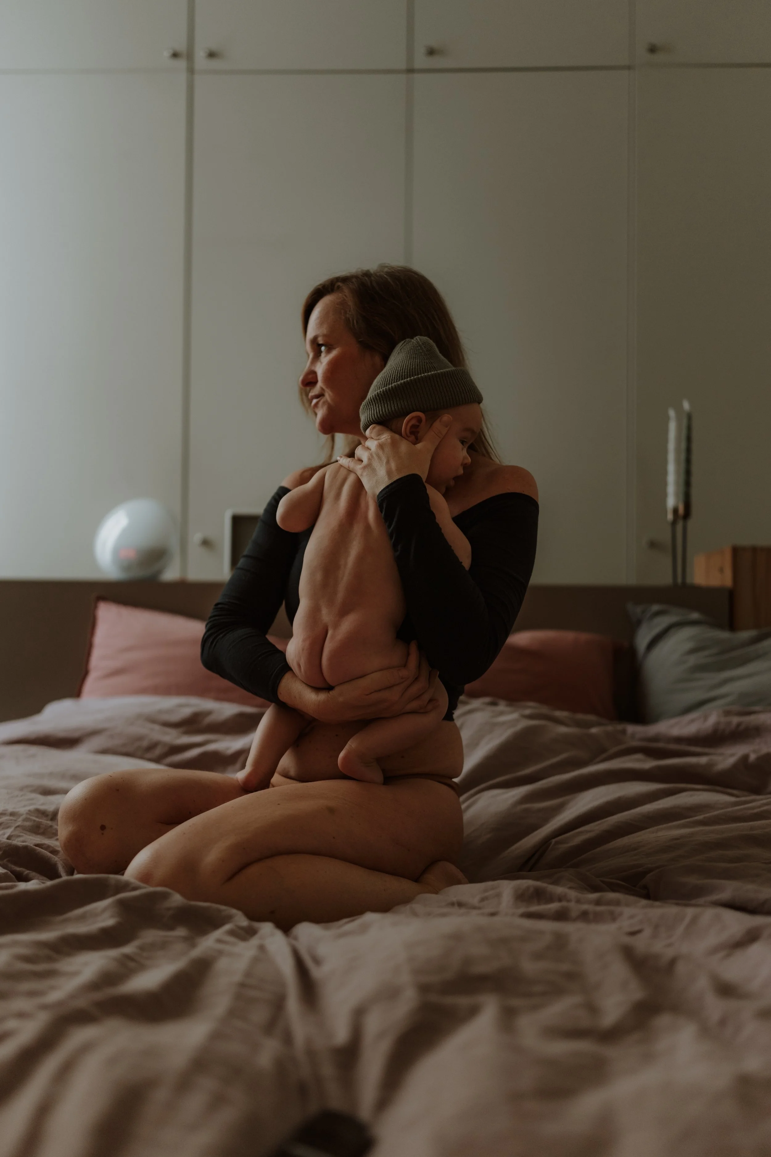 A woman and a young child sitting on a bed in a dimly lit room, with the woman holding the child close. The woman is wearing a black off-shoulder top, and the child is wearing a gray beanie hat and is shirtless.