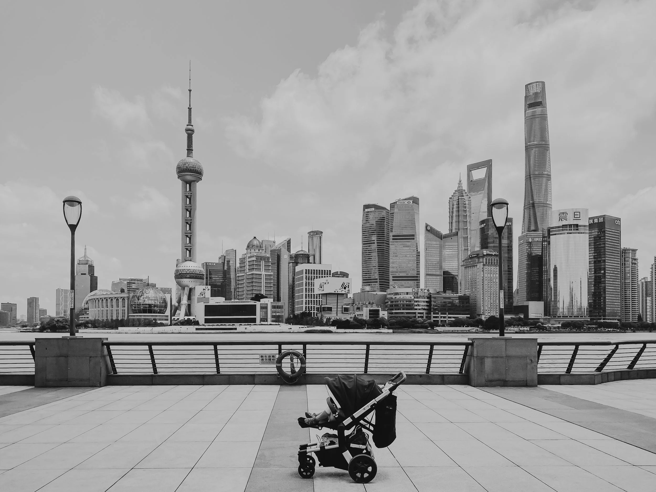Black and white photo of a city skyline with tall skyscrapers, including the Oriental Pearl Tower, across a river. In the foreground, a stroller is parked on a paved promenade with few street lamps and a safety ring on the railing.